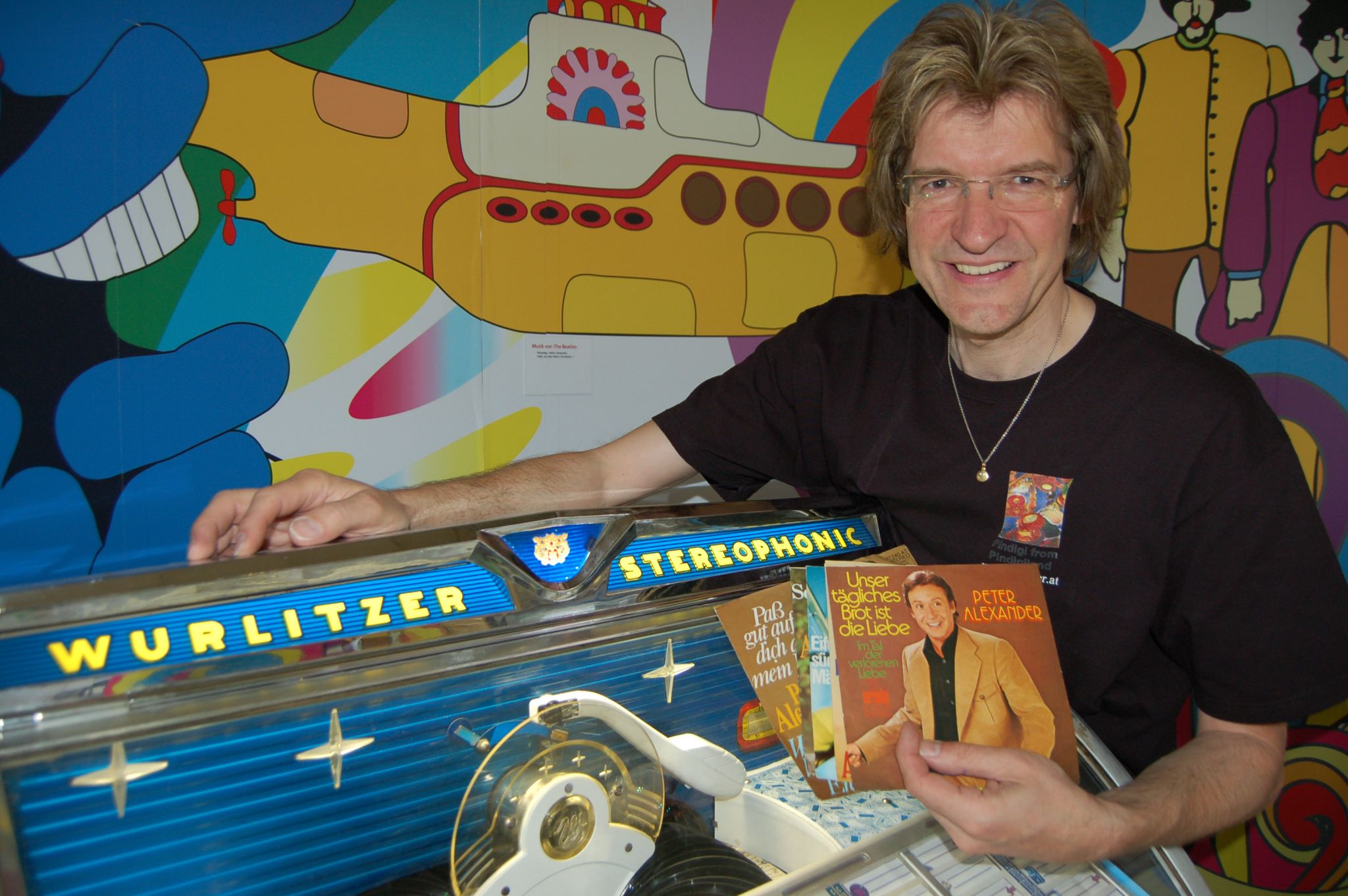 Mann neben einer Wurlitzer-Jukebox mit Schallplatten in der Hand, vor einem bunten Hintergrund mit einem gelben U-Boot.