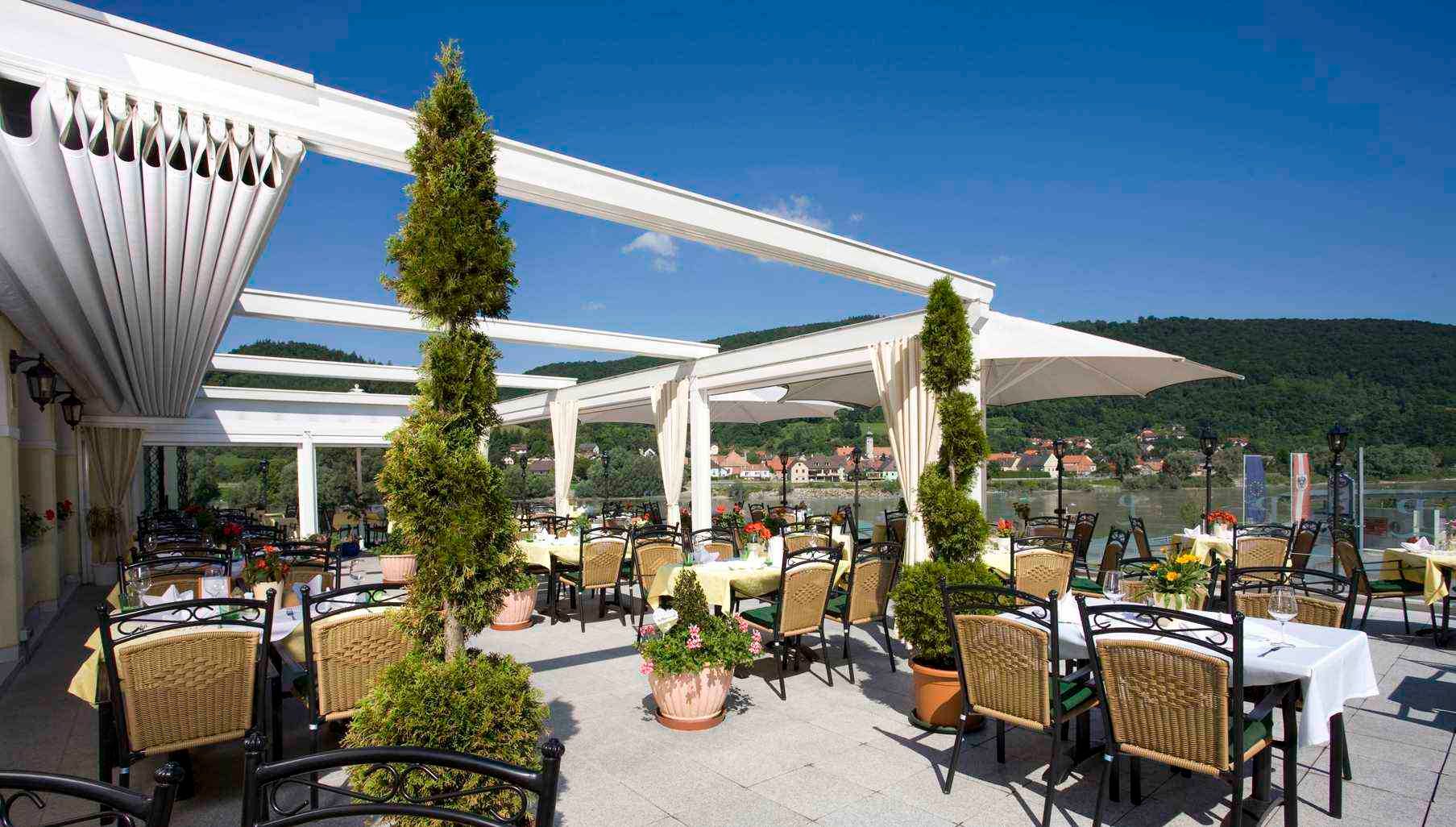Terrasse der Residenz Wachau mit Tischen und Stühlen, umgeben von Pflanzen, mit Blick auf einen Fluss und grüne Hügel im Hintergrund.