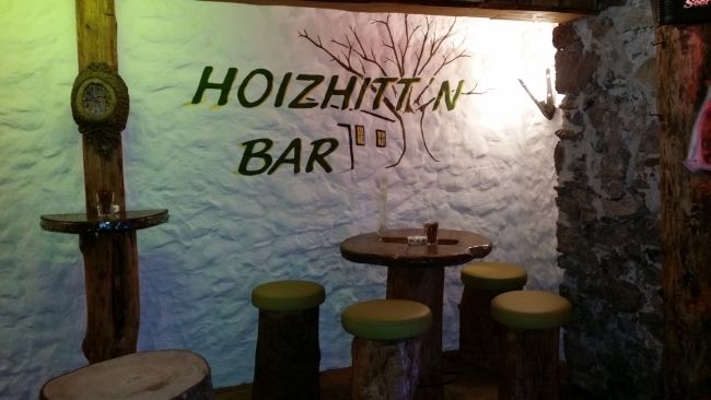 Rustikale Bar mit Holzmöbeln und Schriftzug 'Hoizhitt'n Bar' an der Wand.