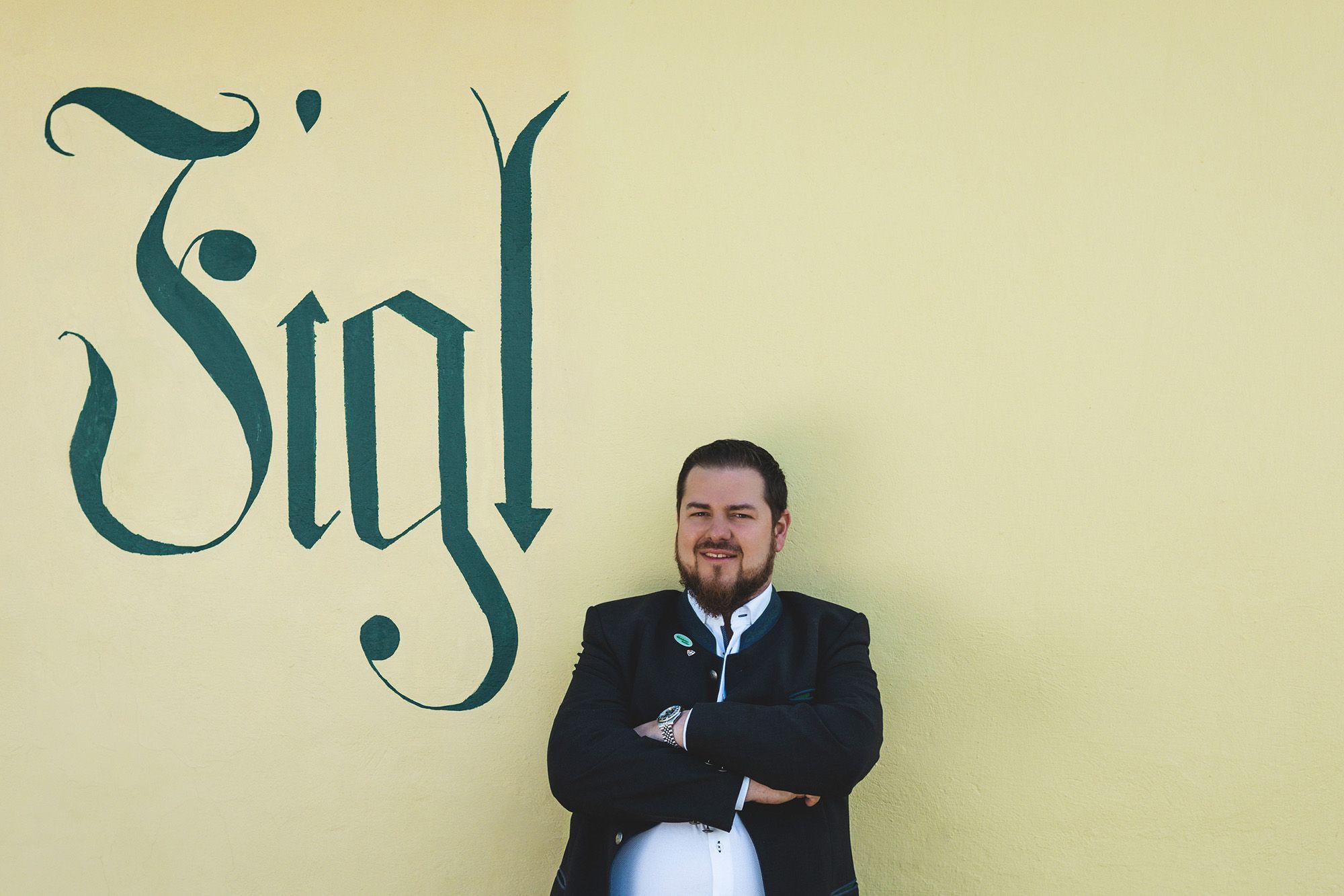 Ein Mann in traditioneller Kleidung steht vor einer gelben Wand mit dem Schriftzug 'Figl'.