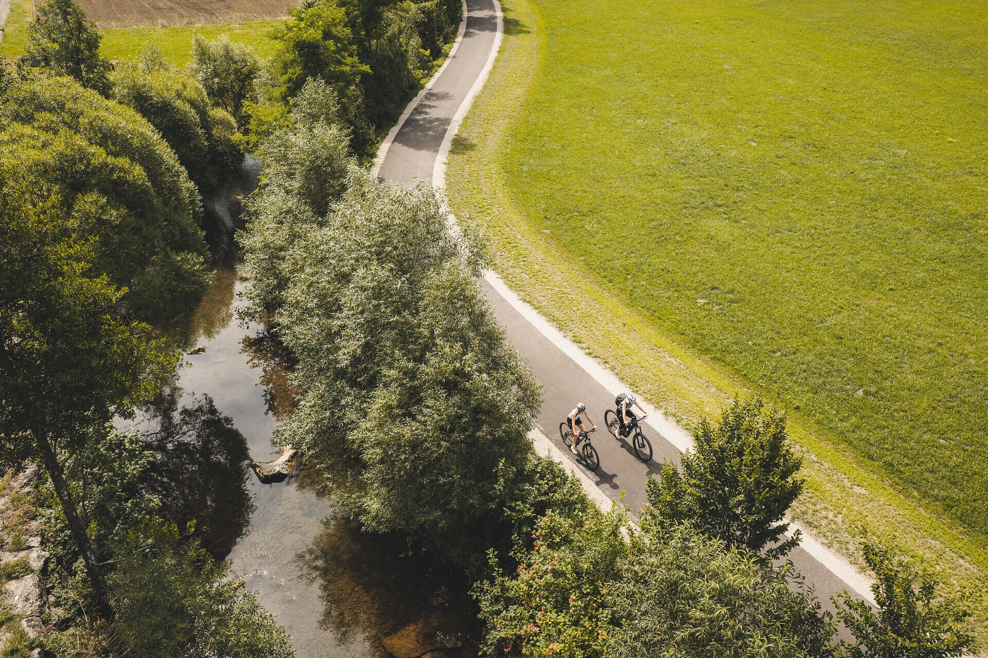 Luftaufnahme von zwei Radfahrerinnen auf einer Straße zwischen Wiesen und Bäumen.