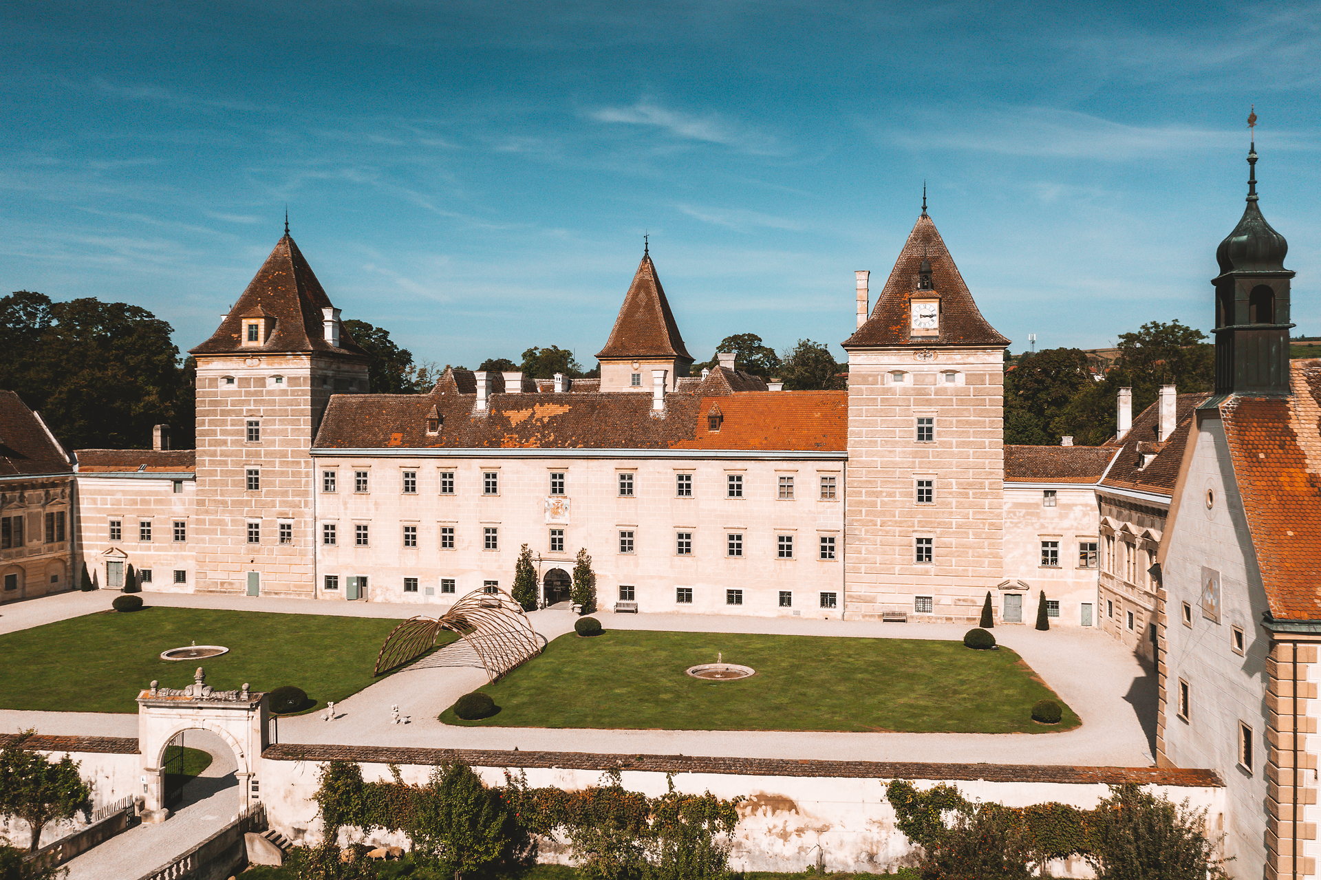 Die majestätischen Türme des Schlosses erheben sich stolz gegen den klaren blauen Himmel. Im weitläufigen Innenhof laden gepflegte Grünflächen und kunstvoll gestaltete Beete zum Verweilen ein. Hier spürt man die harmonische Verbindung von Geschichte und Natur, die jeden Besucher in ihren Bann zieht.