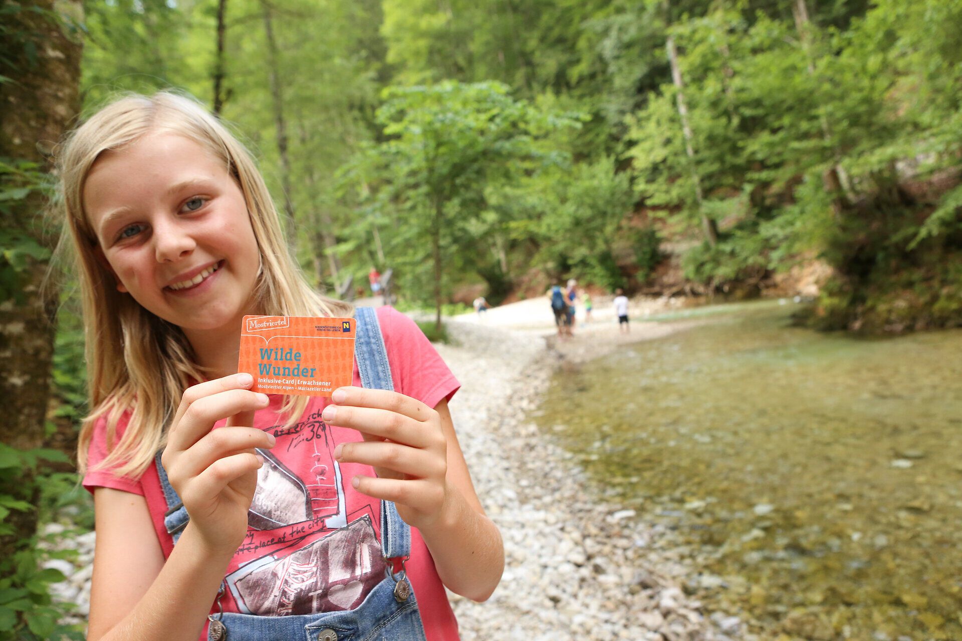 Ein fröhliches Mädchen hält stolz die Wilde Wunder Card in der Hand, während sie am Ufer eines glitzernden Baches steht. Umgeben von üppigem Grün und hohen Bäumen, strahlt die Szene pure Lebensfreude und Abenteuerlust aus. Diese Erlebniskarte verspricht unvergessliche Momente in der Natur.