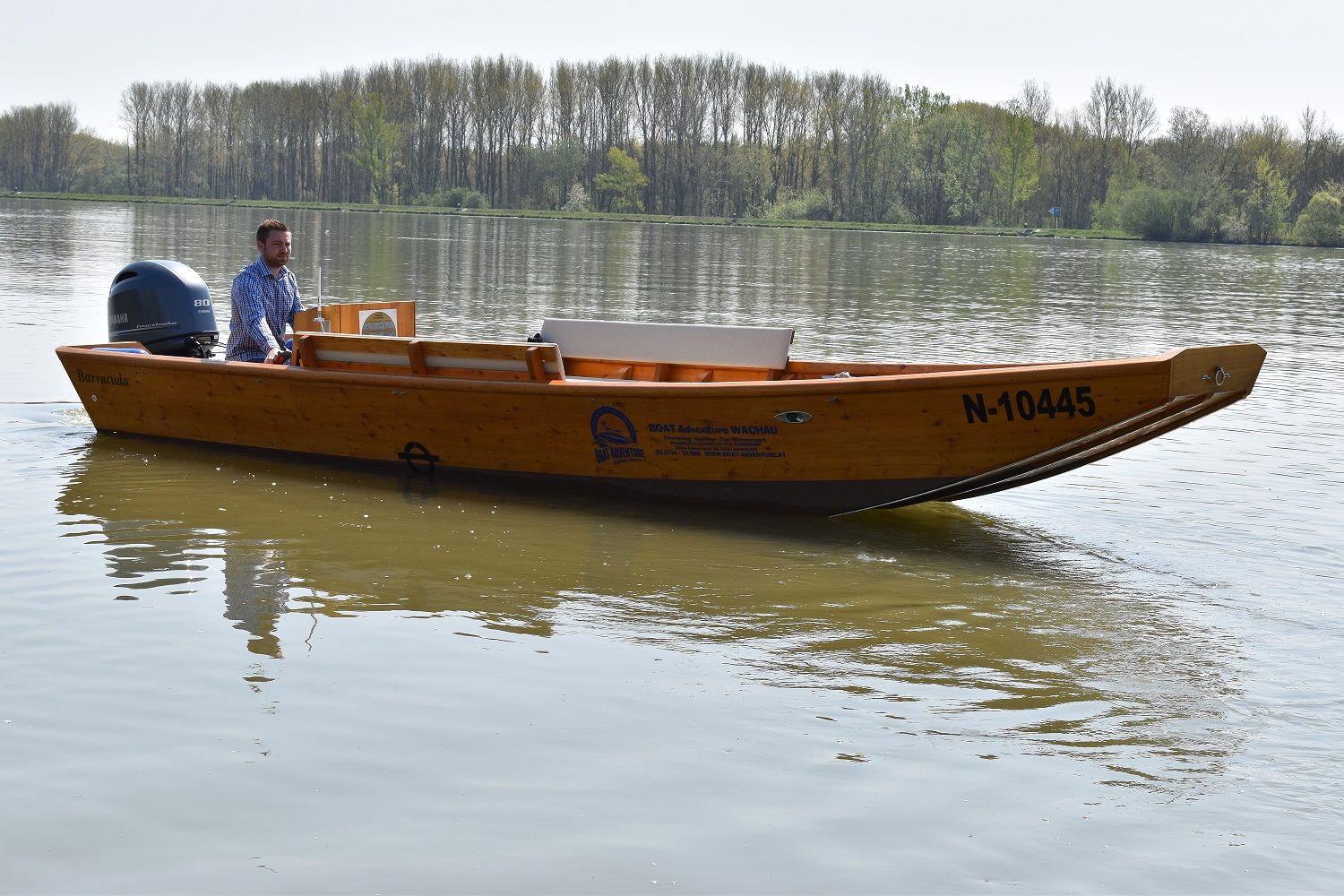 Ein Holzboot mit der Aufschrift 'Barracuda' fährt auf einem ruhigen Fluss. Ein Mann steuert das Boot. Im Hintergrund sind Bäume zu sehen.