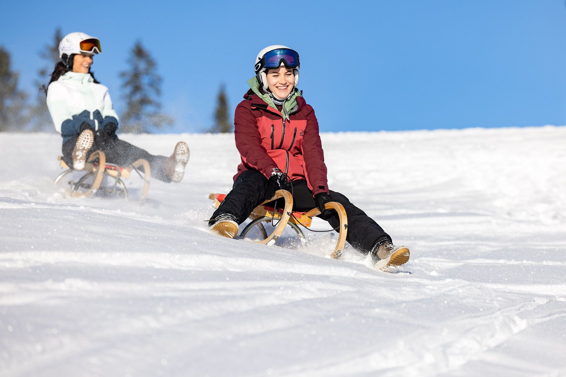 Die strahlende Wintersonne taucht die verschneite Landschaft in ein warmes Licht, während fröhliche Jugendliche mit ihren Schlitten den Hang hinunter sausen. Lachen und Freude erfüllen die Luft, während die schneebedeckten Bäume eine malerische Kulisse bieten. Ein unvergessliches Erlebnis für alle, die den Winter in seiner schönsten Form genießen möchten.