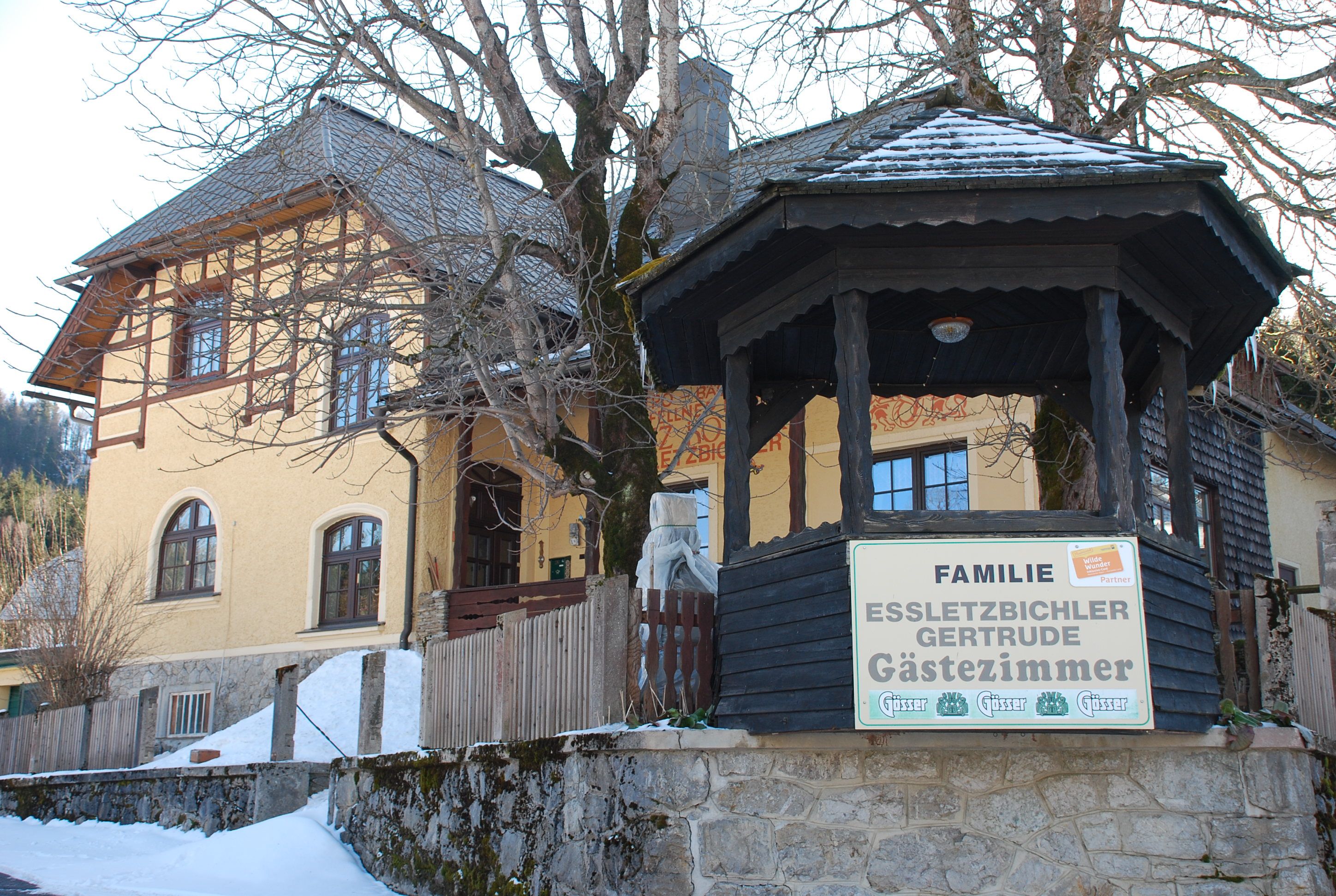 Gelbes Gästehaus mit Holzveranda und Schild 'Familie Essletzbichler Gertrude Gästezimmer'.