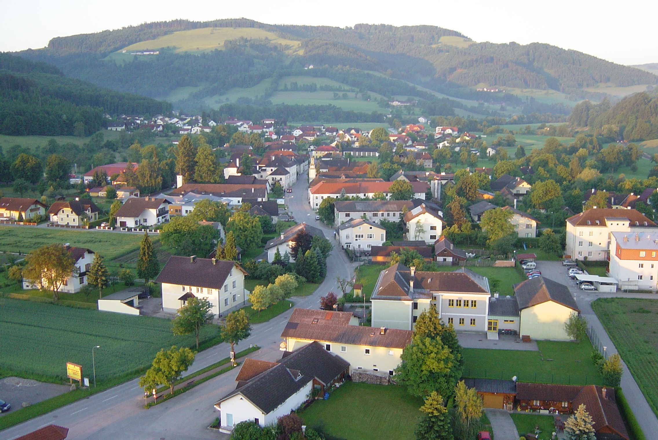 Luftaufnahme eines Dorfes mit Häusern, Straßen und umliegenden Hügeln im Hintergrund.