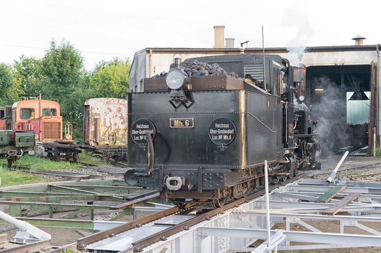 Dampflokomotive vor einem Heizhaus mit Schienen im Vordergrund.