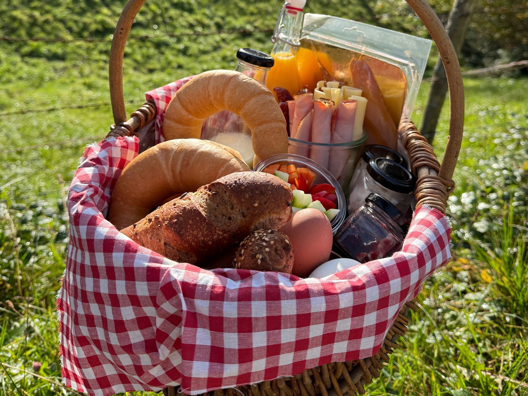Ein Picknickkorb mit Brot, Croissants, Eiern, Saft, Aufschnitt und Gemüse auf einer rot-weiß karierten Decke im Freien.