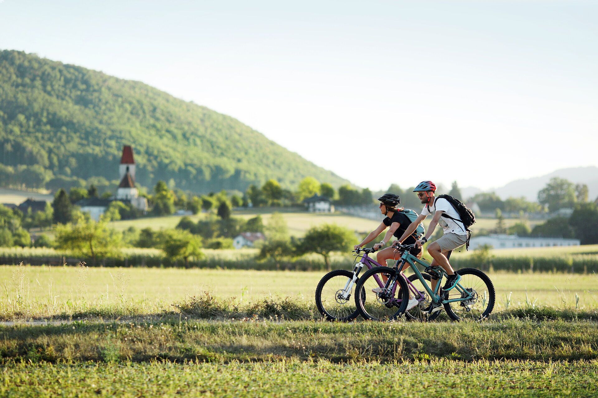 Pärchen beim Radfahren, im Hintergrund ist eine Kirche zu sehen.