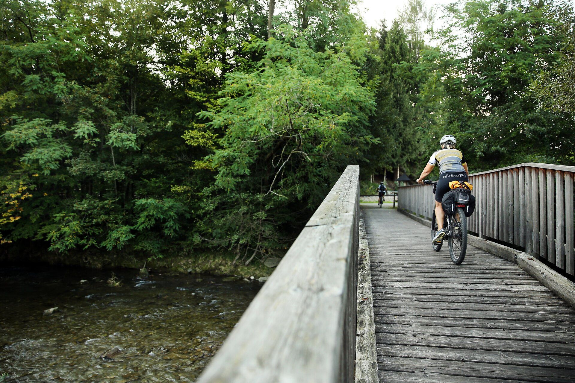 Eine malerische Brücke führt Radfahrer über einen sanft plätschernden Fluss, umgeben von üppigem Grün und der Ruhe der Natur. Die frische Luft und das sanfte Licht schaffen eine einladende Atmosphäre für alle, die die Schönheit der Umgebung genießen möchten.