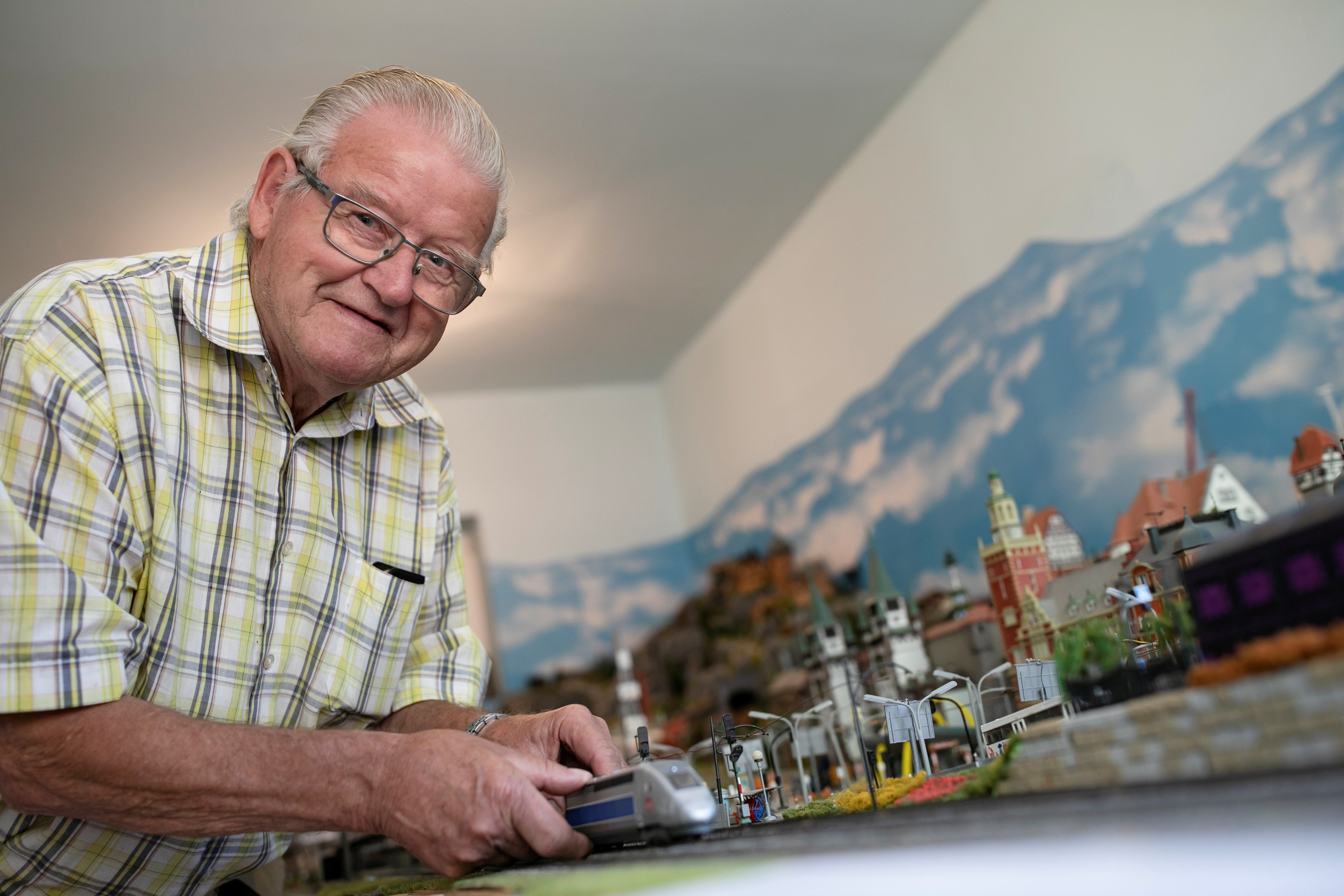 Opa Moser steht vor seiner Modelleisenbahnlandschaft und zeigt eine Lokomotive. 