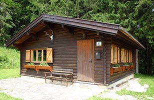Ein kleines, rustikales Jagdhaus aus Holz mit Fensterläden und Geweih an der Fassade, umgeben von Wald.