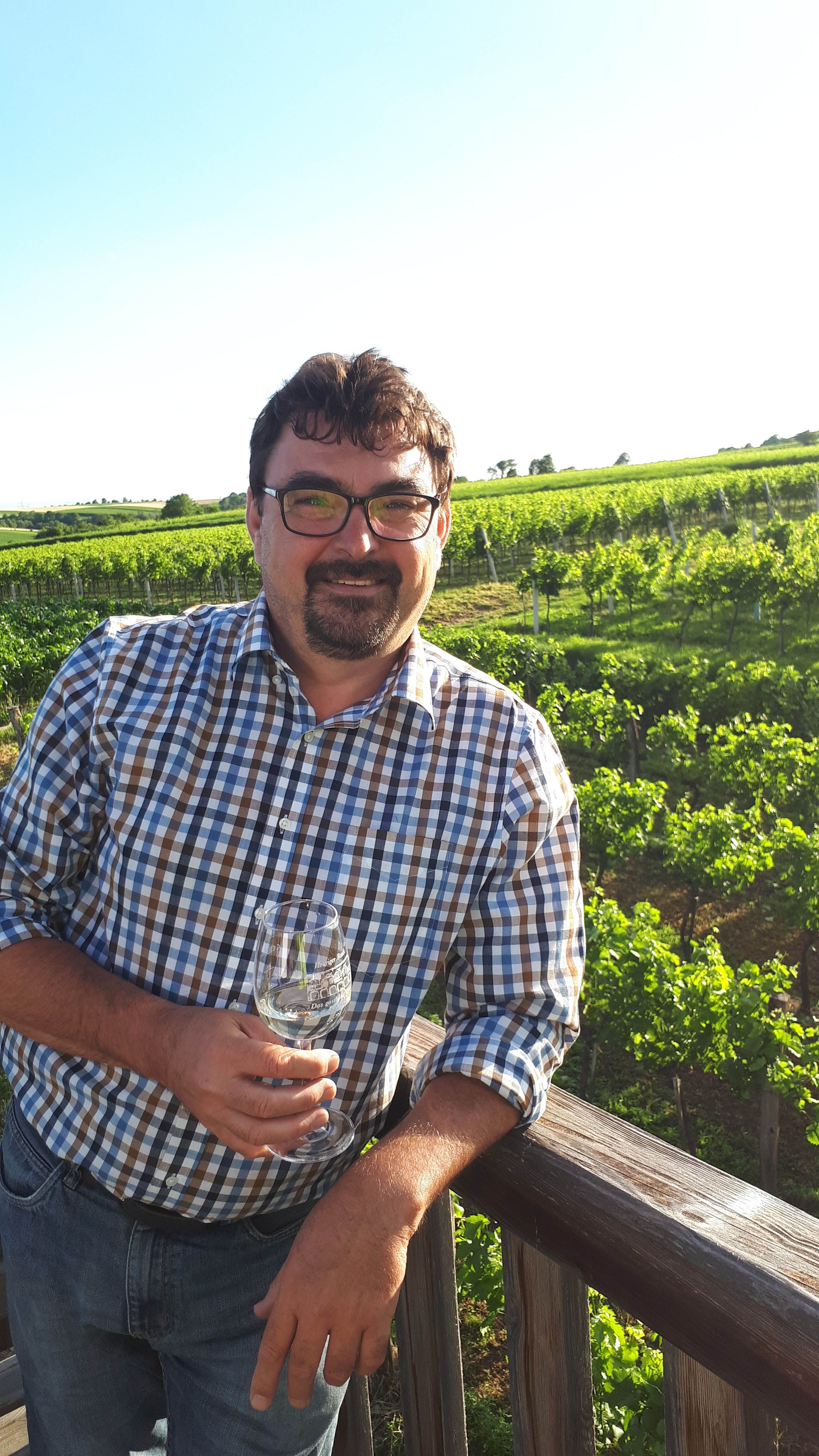 Ein Mann im karierten Hemd steht mit einem Weinglas in der Hand vor einem Weinberg.