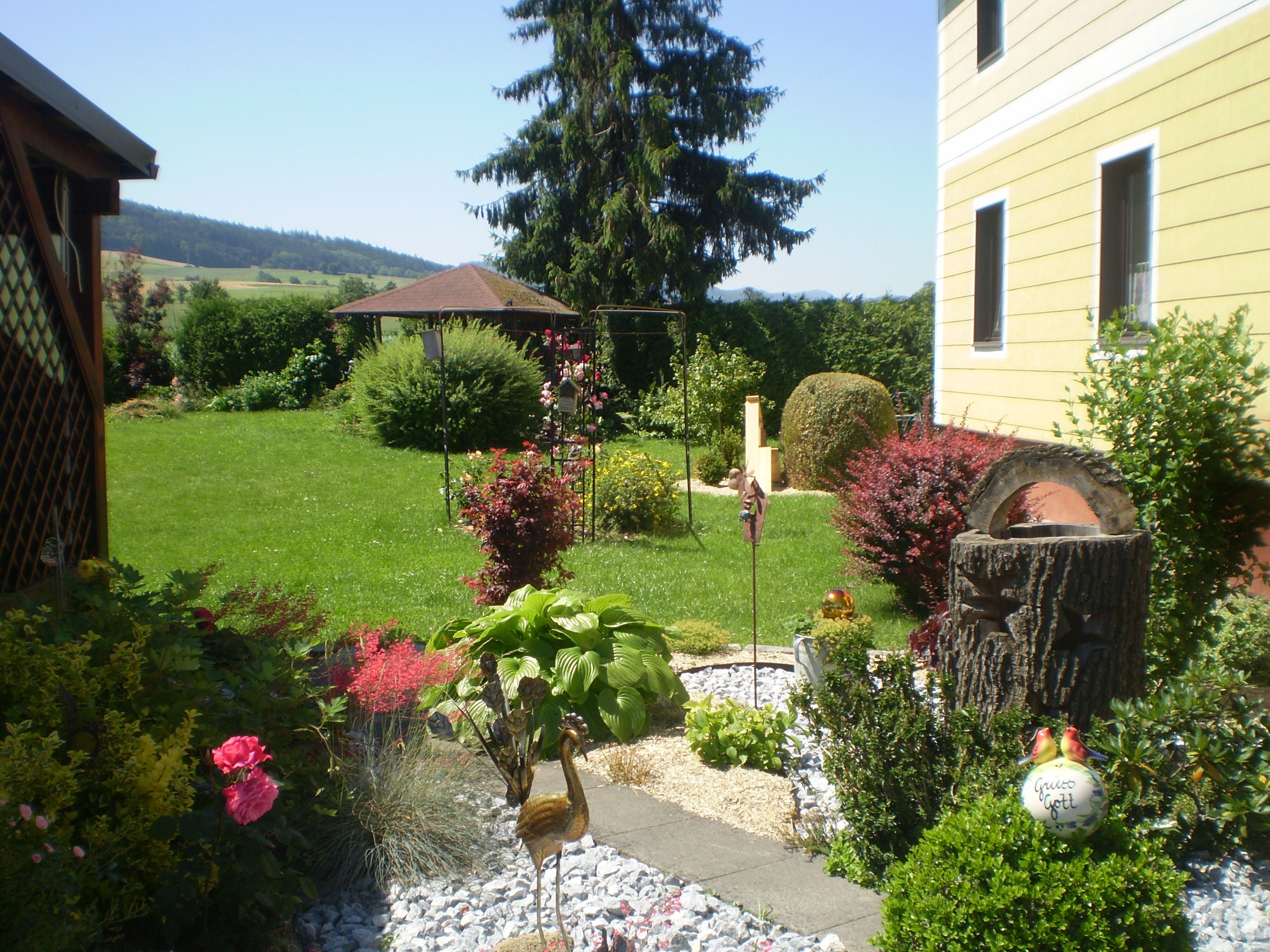 Ein gepflegter Garten mit Blumen, Sträuchern und einem gelben Haus im Hintergrund.