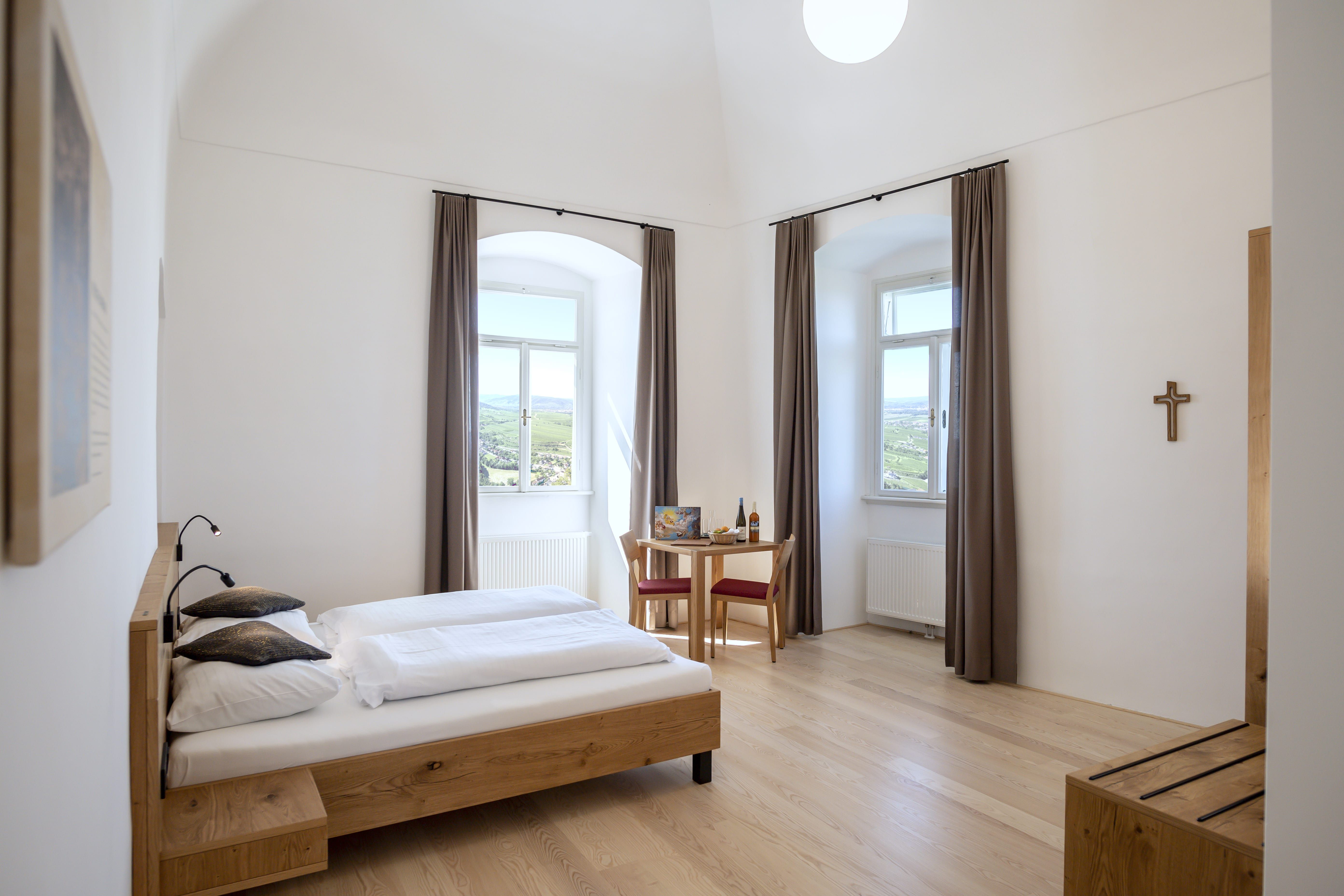 Helles Doppelzimmer mit Holzbett, Tisch und Stühlen, zwei Fenstern mit Vorhängen und einem Kreuz an der Wand.