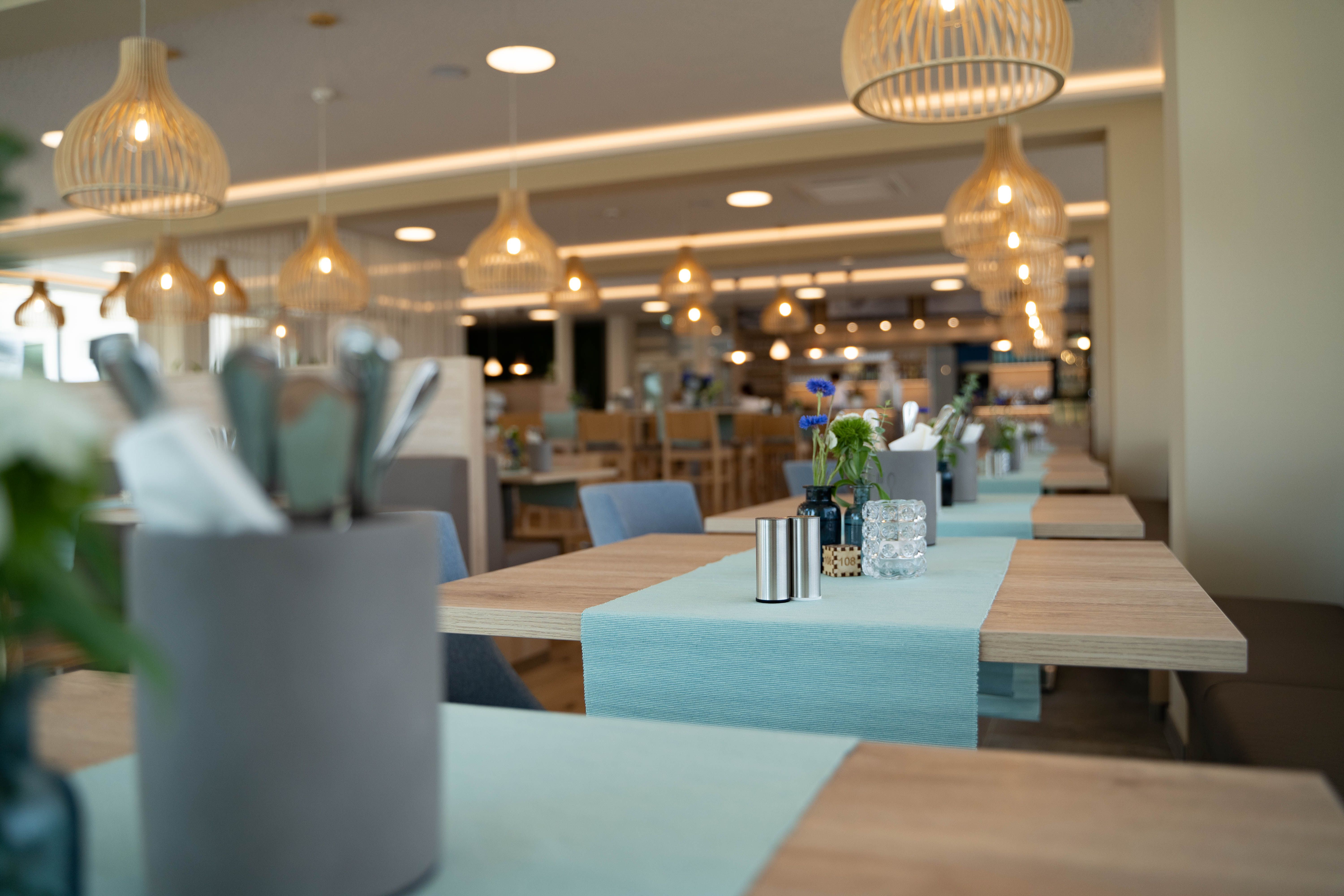 Innenansicht eines modernen Cafés mit Holztischen, blauen Tischläufern und hängenden Lampen.