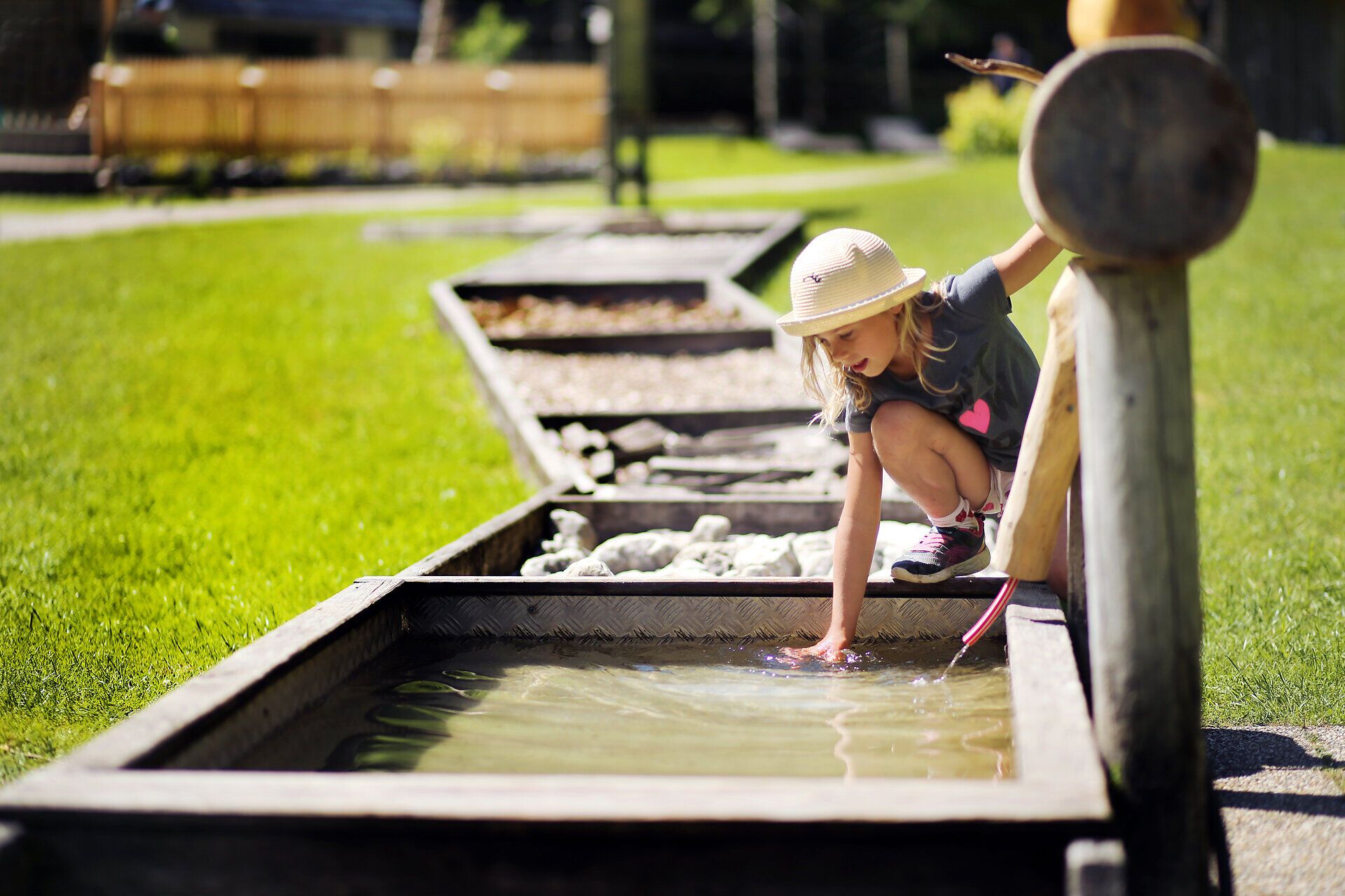 Ein fröhliches Mädchen taucht ihre Hand ins kühle Wasser eines kleinen Baches, während die Sonne sanft auf die grüne Wiese scheint. Umgeben von der idyllischen Natur des Mendlingtals, strahlt die Szene eine unbeschwerte Sommerfreude aus, die zum Spielen und Entdecken einlädt.