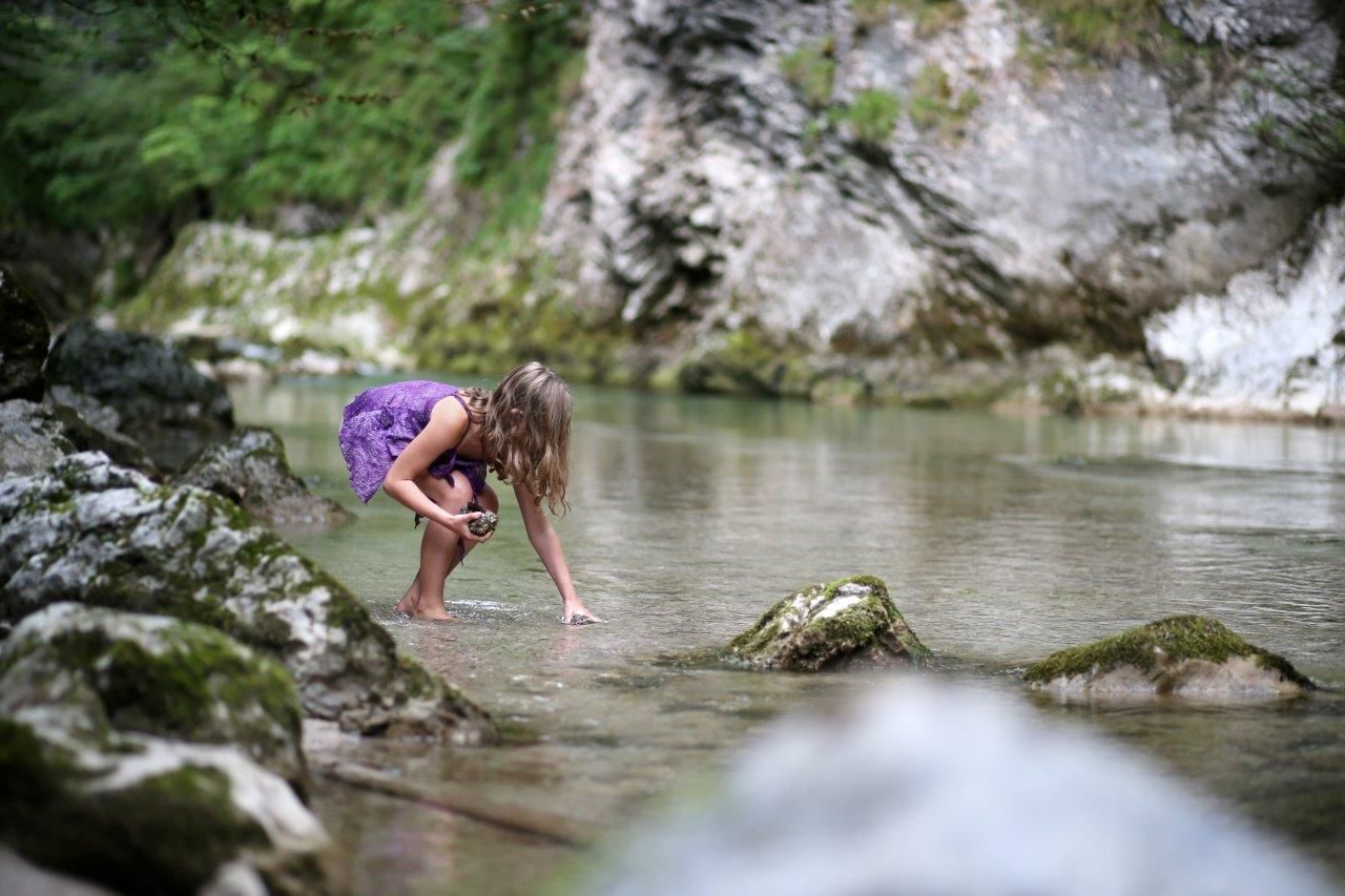 Ein Kind im lila Kleid spielt in einem klaren Bach, umgeben von Felsen und grüner Natur.