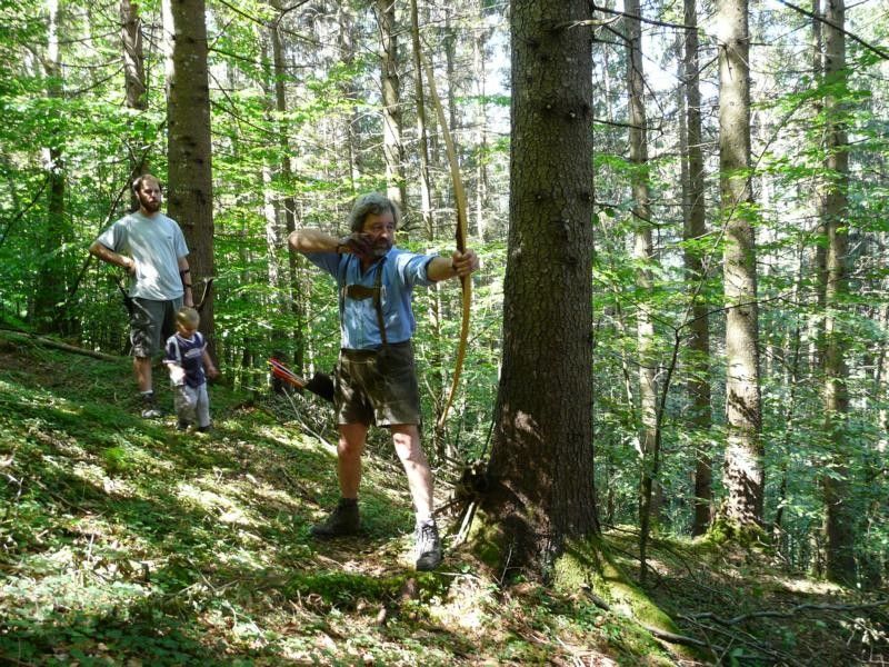 Person schießt mit einem Bogen im Wald, zwei weitere Personen im Hintergrund.