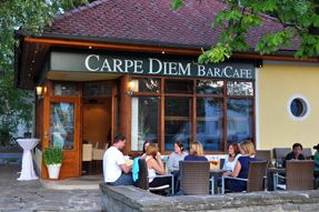 Außenansicht eines Cafés mit dem Namen Carpe Diem, Menschen sitzen an Tischen im Freien.