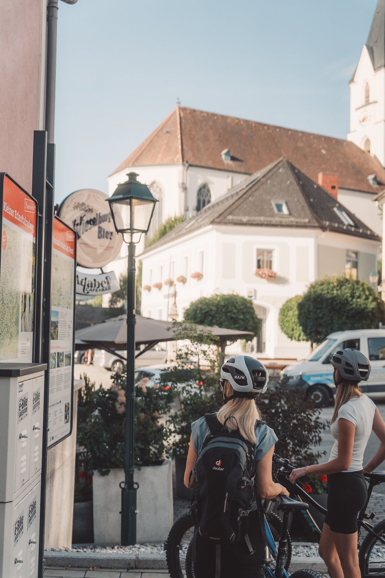 Radfahrerinnen betrachten eine Infotafel, im Hintergrund eine Kirche.
