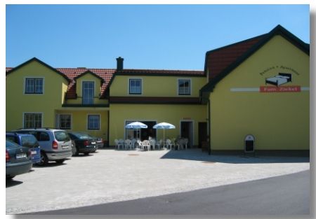 Gelbes Gebäude mit der Aufschrift 'Pension Ziekel', Autos und Tische mit Sonnenschirmen davor.