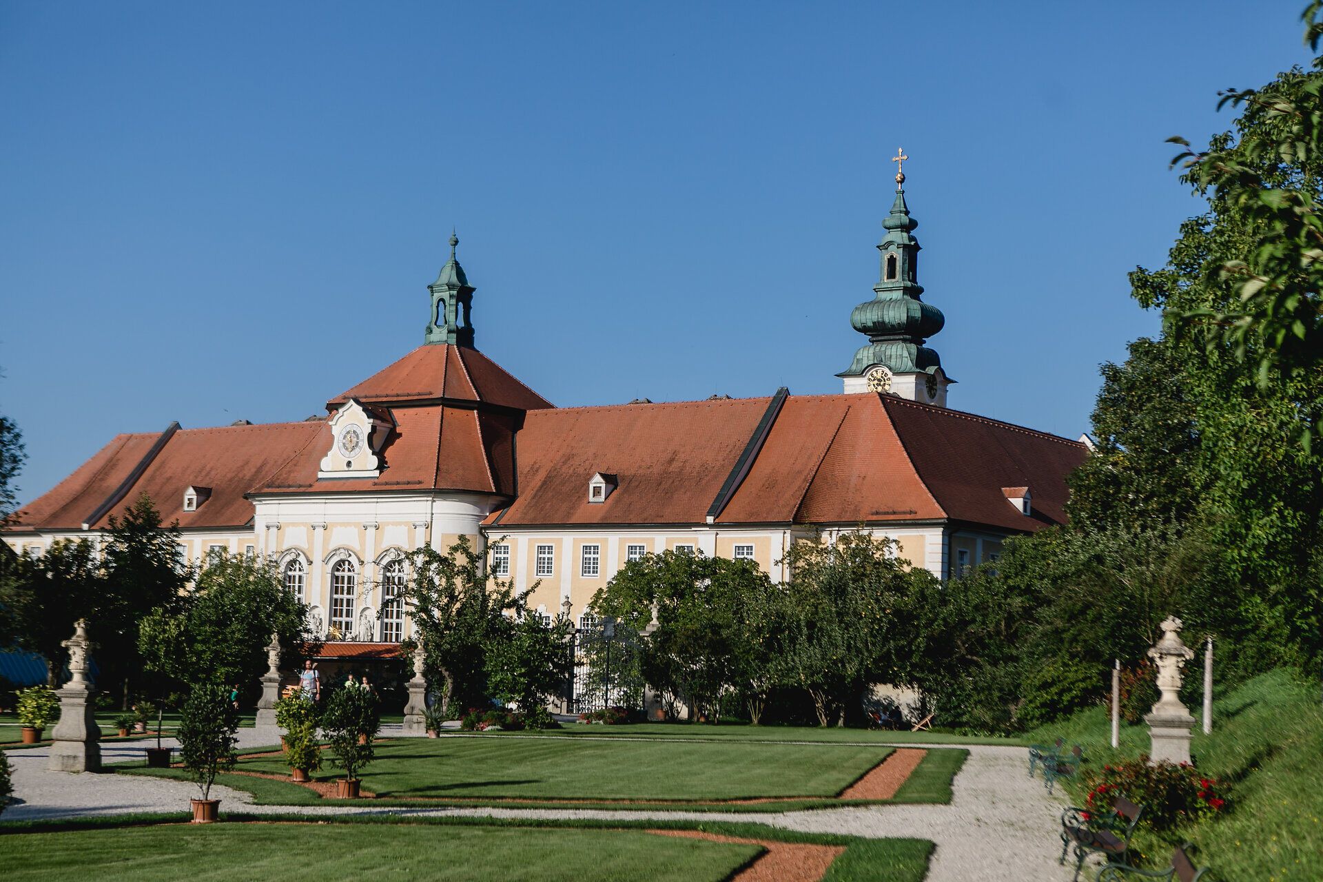 Der historische Hofgarten des Stifts Seitenstetten lädt mit seinen gepflegten Rasenflächen und blühenden Pflanzen zum Verweilen ein. Die majestätische Architektur des Gebäudes harmoniert perfekt mit der klaren blauen Himmel, während die sanften Hügel im Hintergrund eine malerische Kulisse bieten.