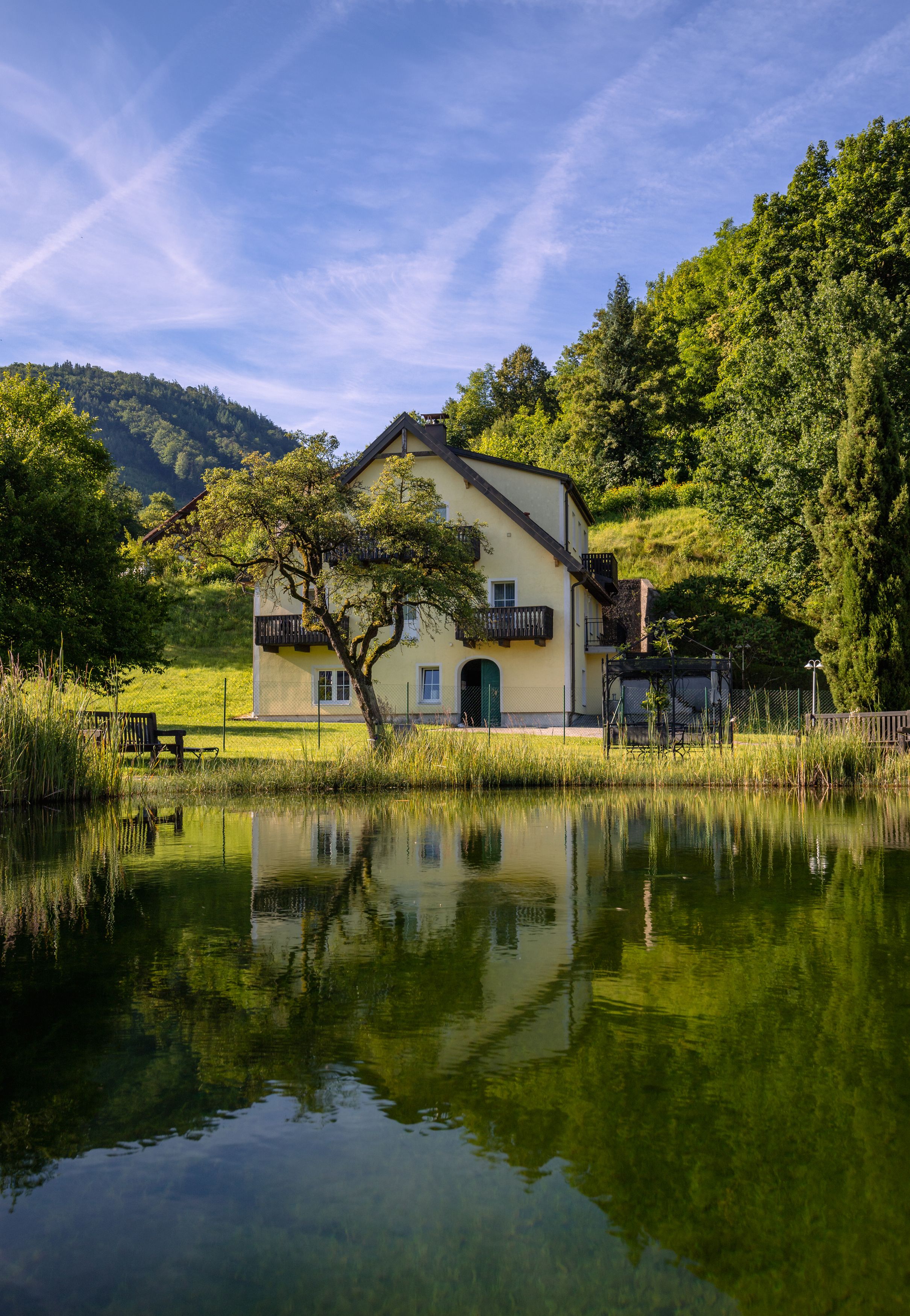 Gelbes Haus mit Balkon, umgeben von Bäumen, spiegelt sich in einem Teich.