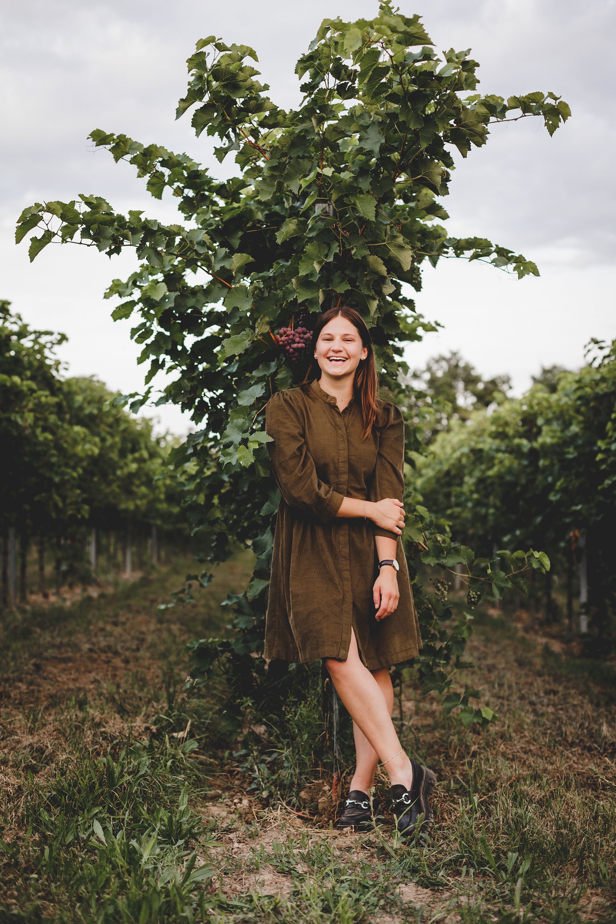 Eine Frau steht lächelnd in einem Weinberg vor einer Weinrebe.
