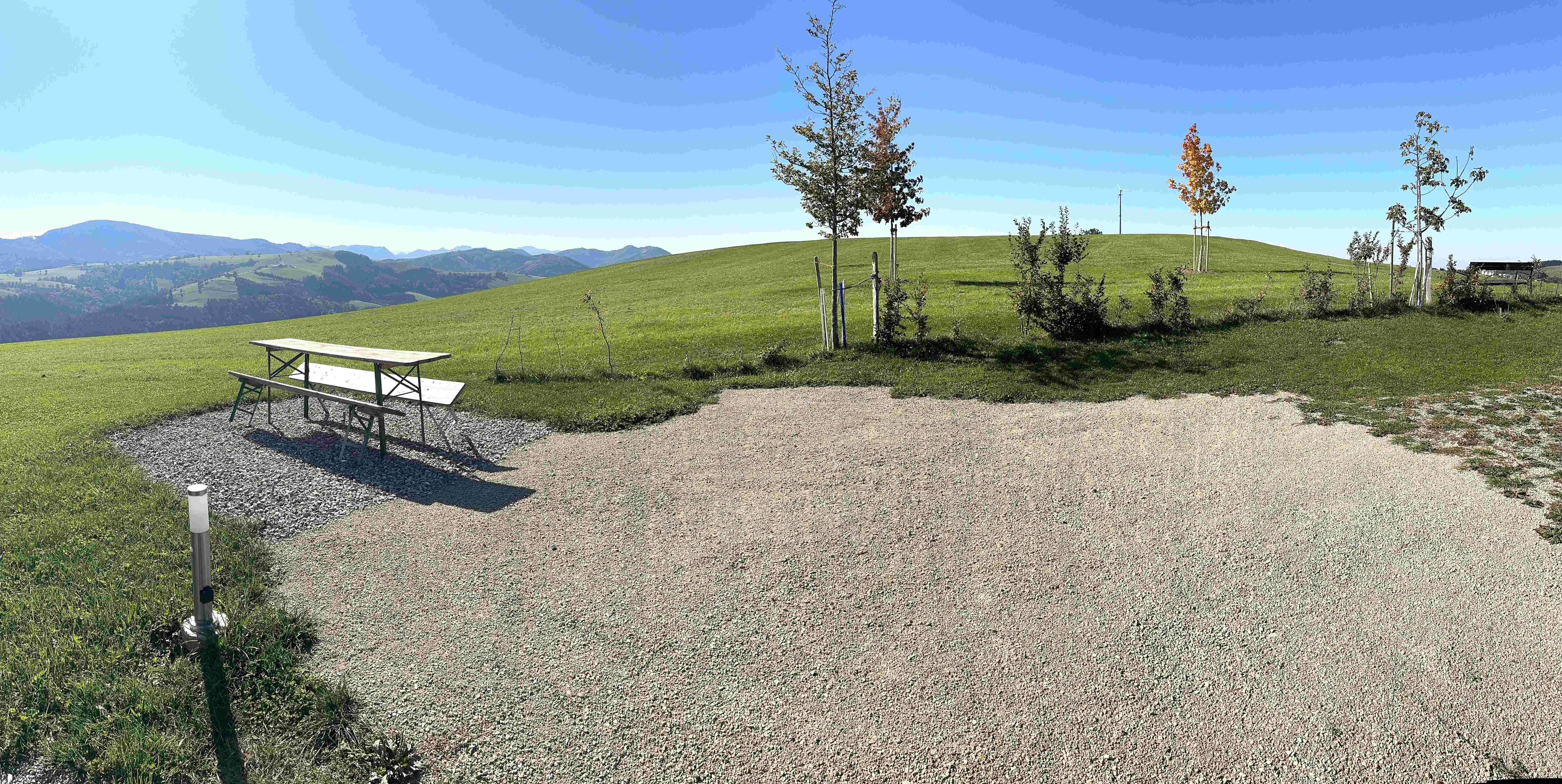 Panorama vom Camping am Hof Rabenberg mit Sitzgarnitur, Kiesflächen, jungen Bäumen und Blick auf die Berge unter blauem Himmel.