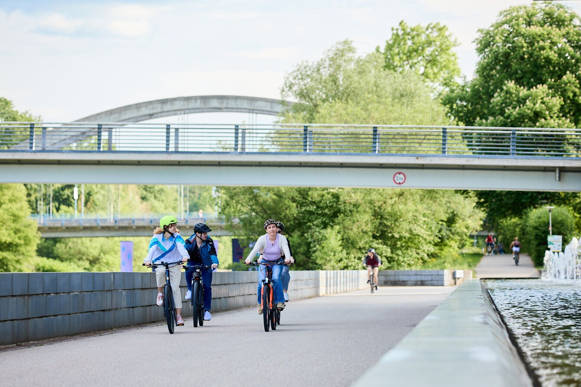 Mehrere Menschen fahren mit dem Rad, im Hintergrund sind eine Brücke, grüne Bäume und weitere Radfahrende bei sonnigem Wetter zu sehen