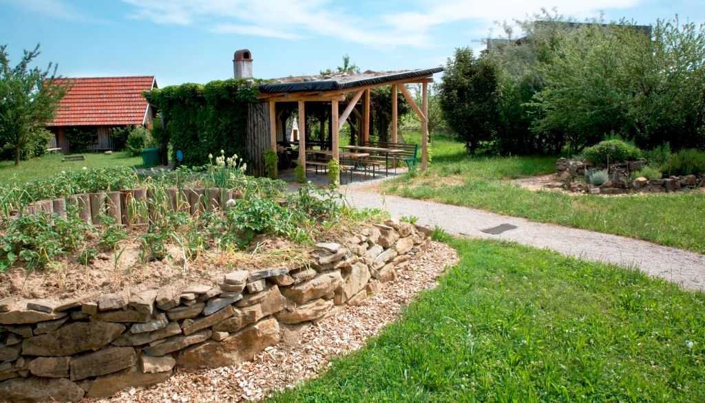 Ein Garten mit Hochbeet aus Steinen, überdachtem Sitzbereich und Rasenfläche.
