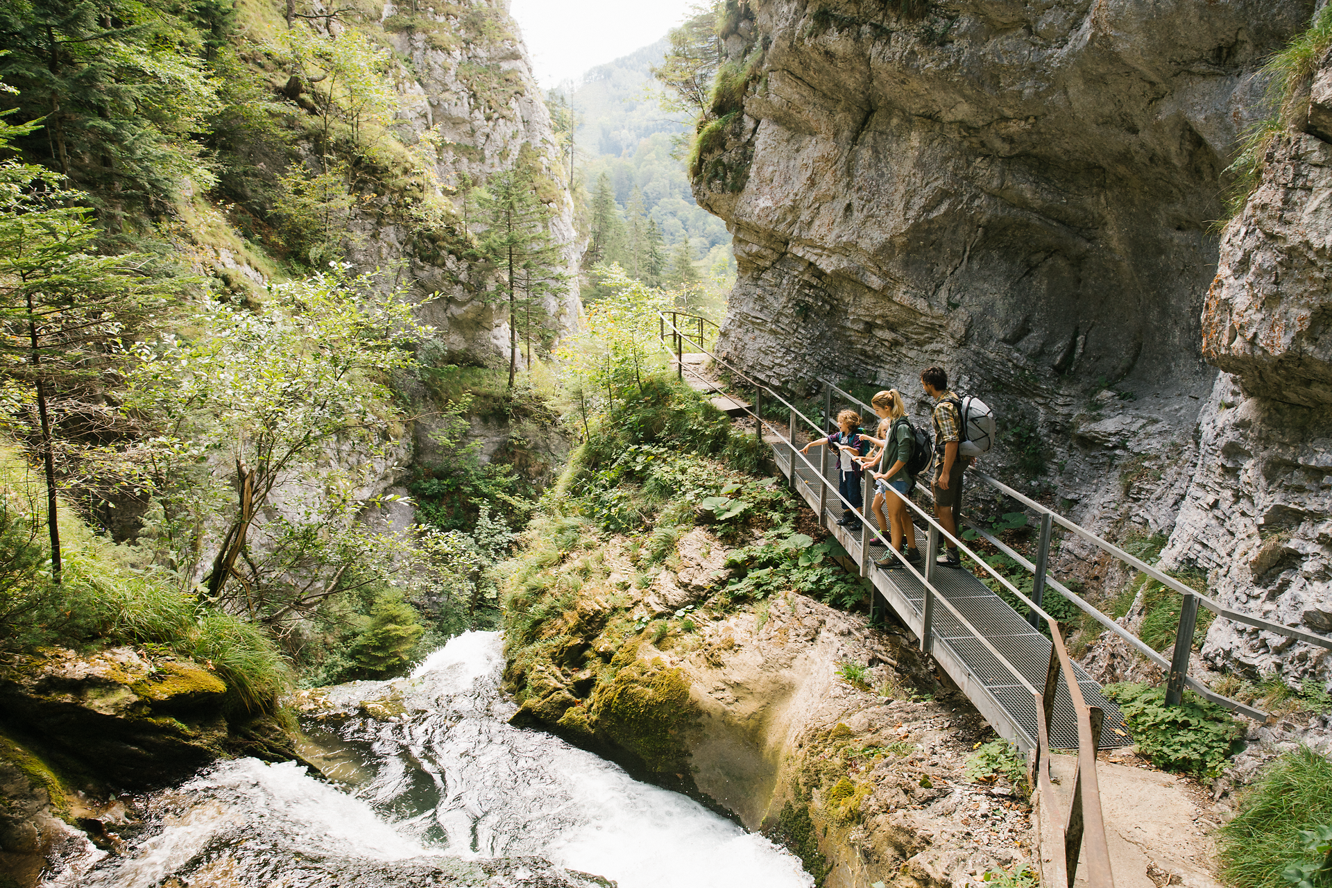 Ein malerischer Wanderweg schlängelt sich entlang des rauschenden Wassers, umgeben von üppigem Grün und majestätischen Felsen. Familien genießen die frische Luft und die atemberaubenden Ausblicke, während sie die Natur in vollen Zügen erleben.