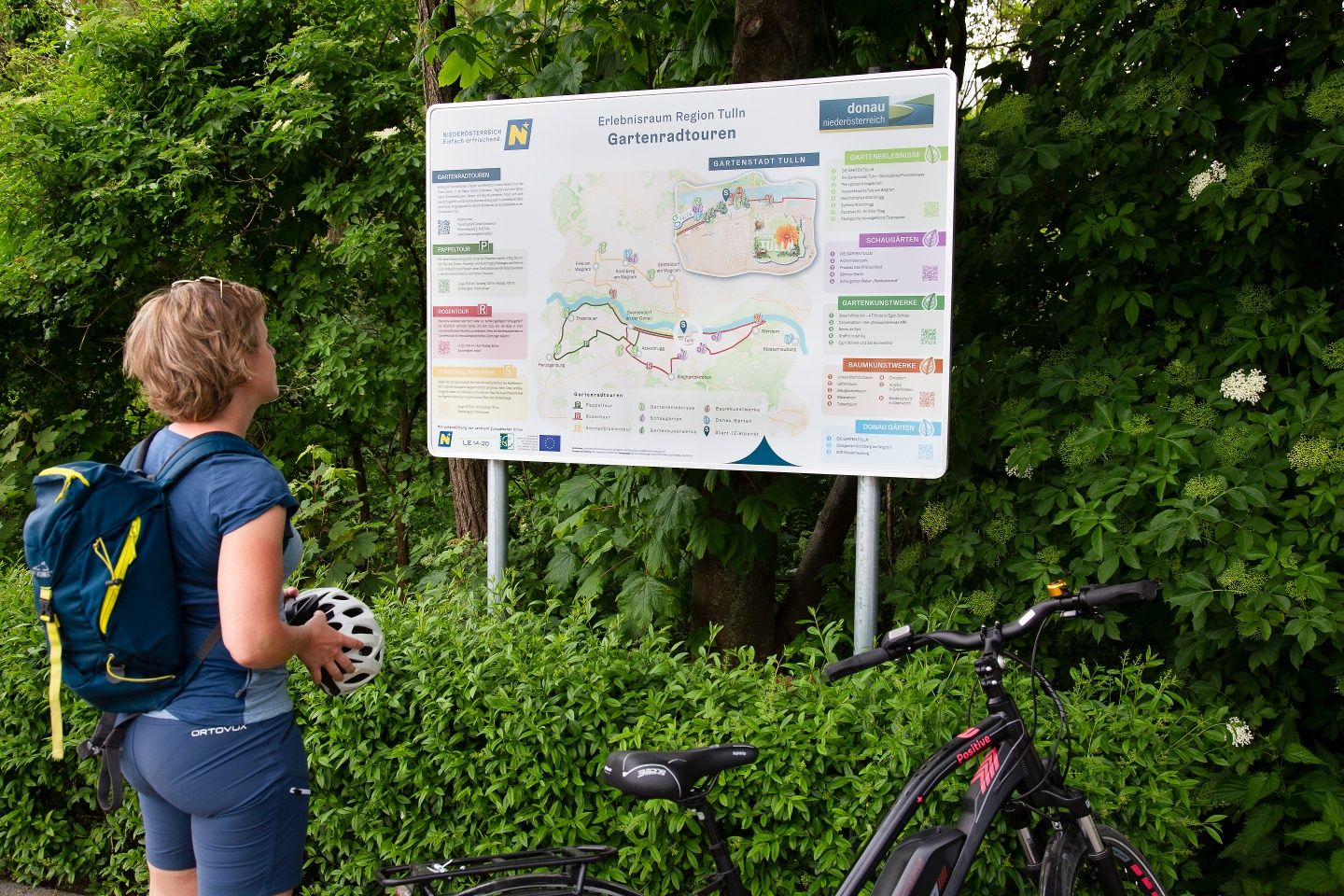 Eine Frau mit Fahrradhelm und Rucksack betrachtet eine Infotafel über Gartenradtouren in der Region Tulln, umgeben von grüner Vegetation.