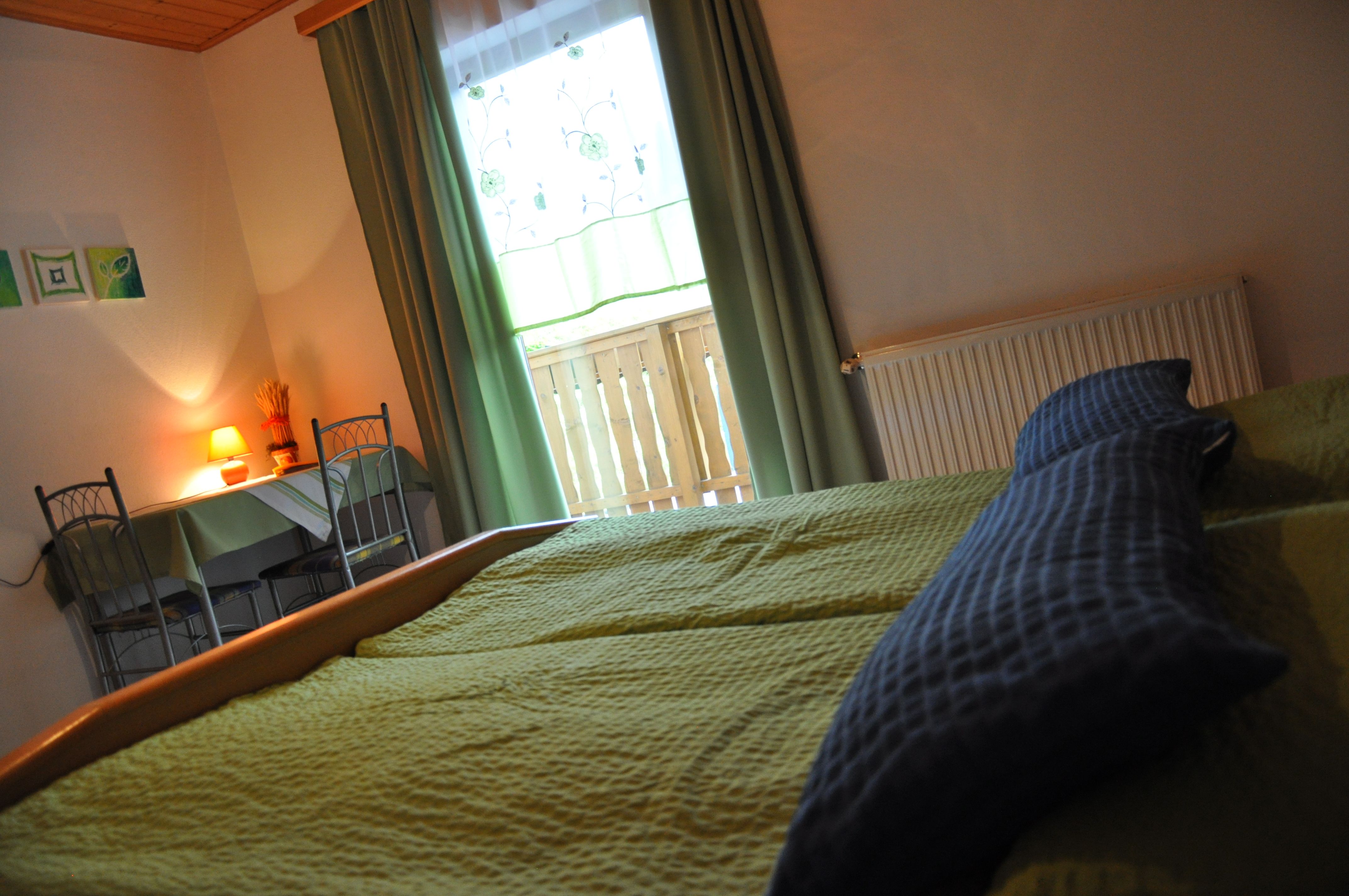 Gemütliches Schlafzimmer mit grünem Bett, Tisch und Stühlen, Lampe und Fenster mit Vorhängen.