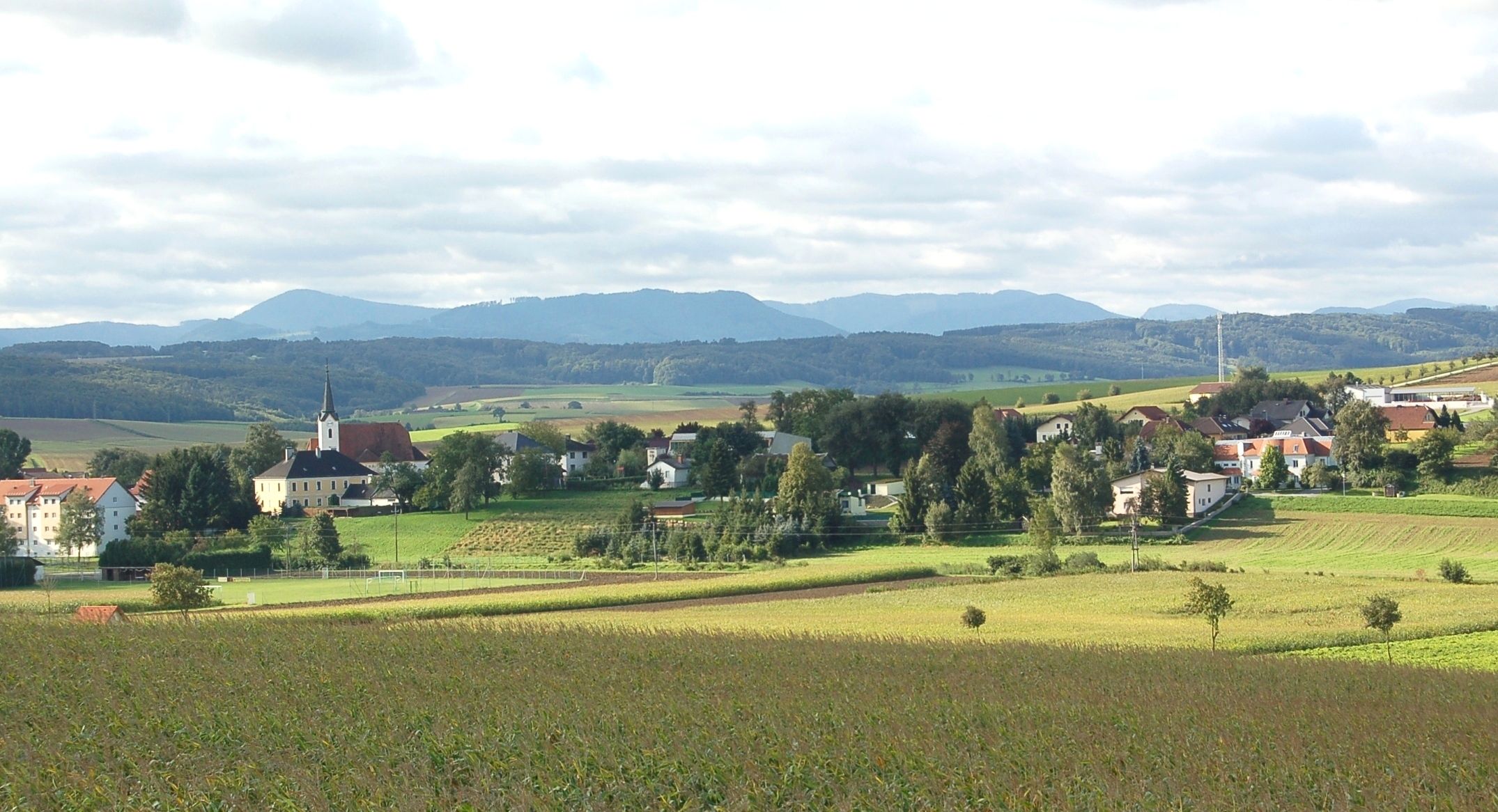 Landschaft mit Dorf und Kirche in St. Margarethen an der Sierning, umgeben von Feldern und Hügeln.