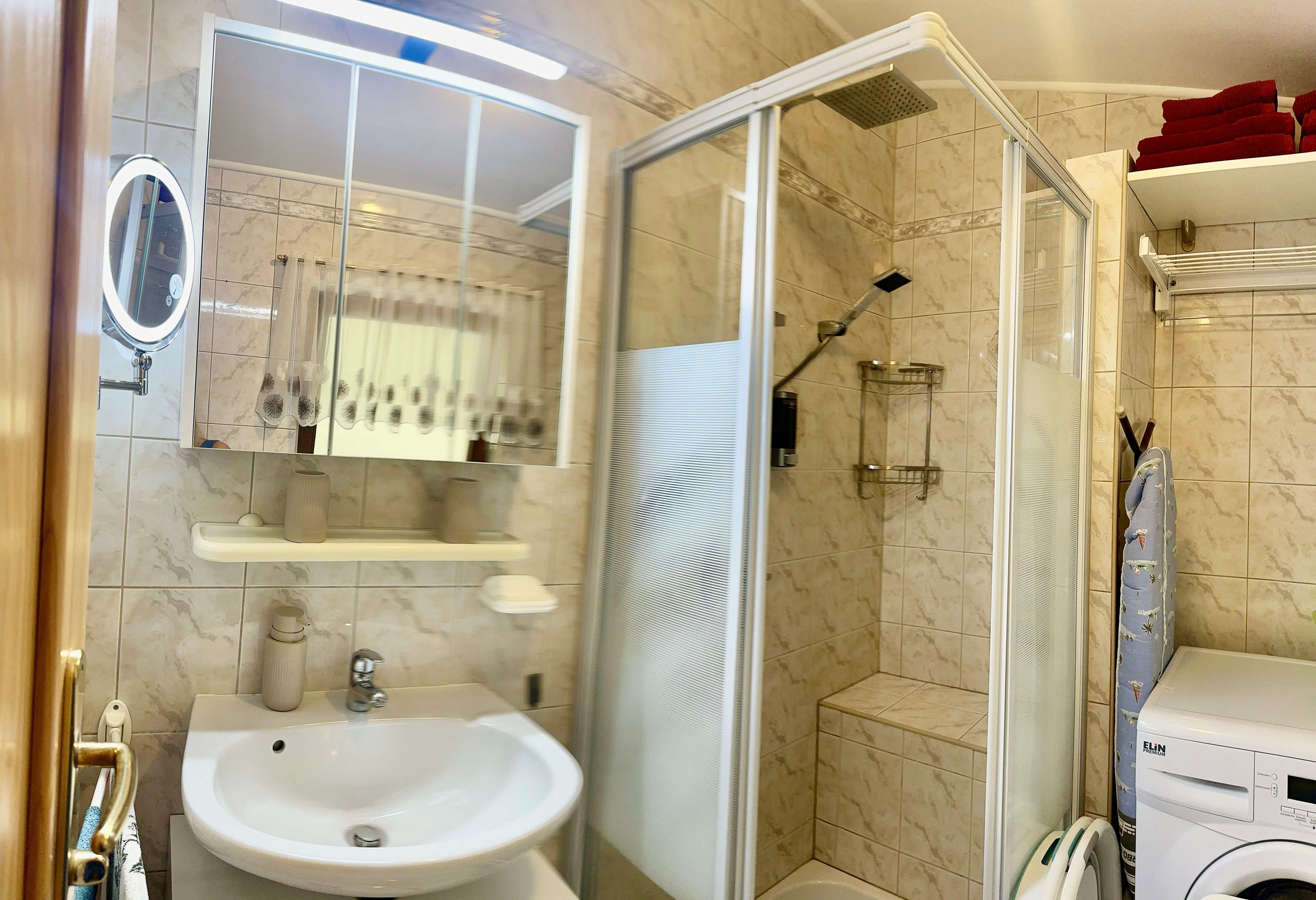 Badezimmer mit Dusche, Waschbecken, Spiegel und Waschmaschine.