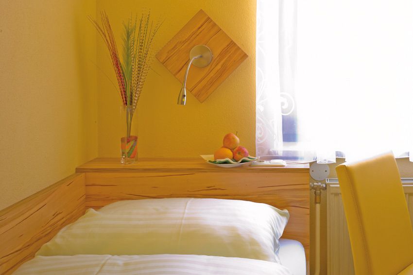 Gemütliches Zimmer mit gelben Wänden, Bett, Vase mit Gräsern und Obstschale auf Holzregal.