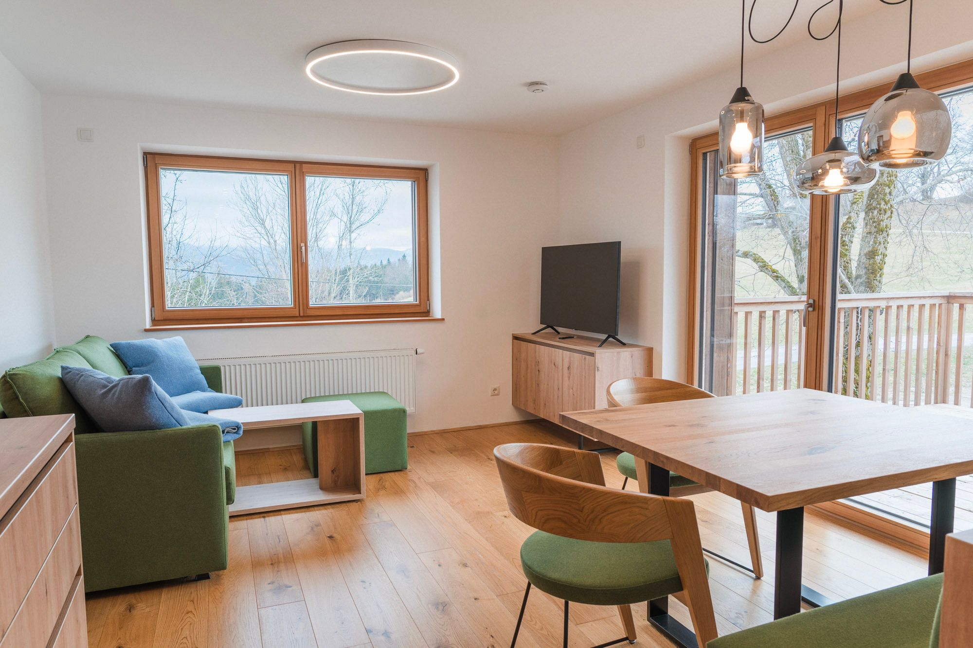 Modern eingerichtetes Wohnzimmer mit Holzboden, grüner Couch, Esstisch und großen Fenstern mit Blick ins Freie.