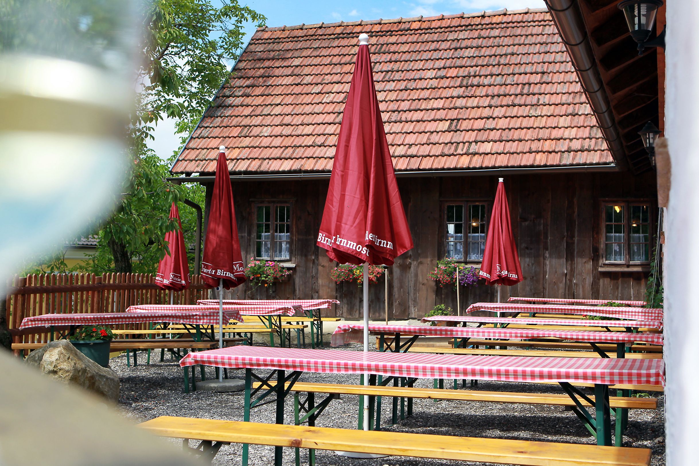 Ein traditioneller Heuriger mit Holztischen, rot-weiß karierten Tischdecken und roten Sonnenschirmen vor einem rustikalen Gebäude mit Ziegeldach.