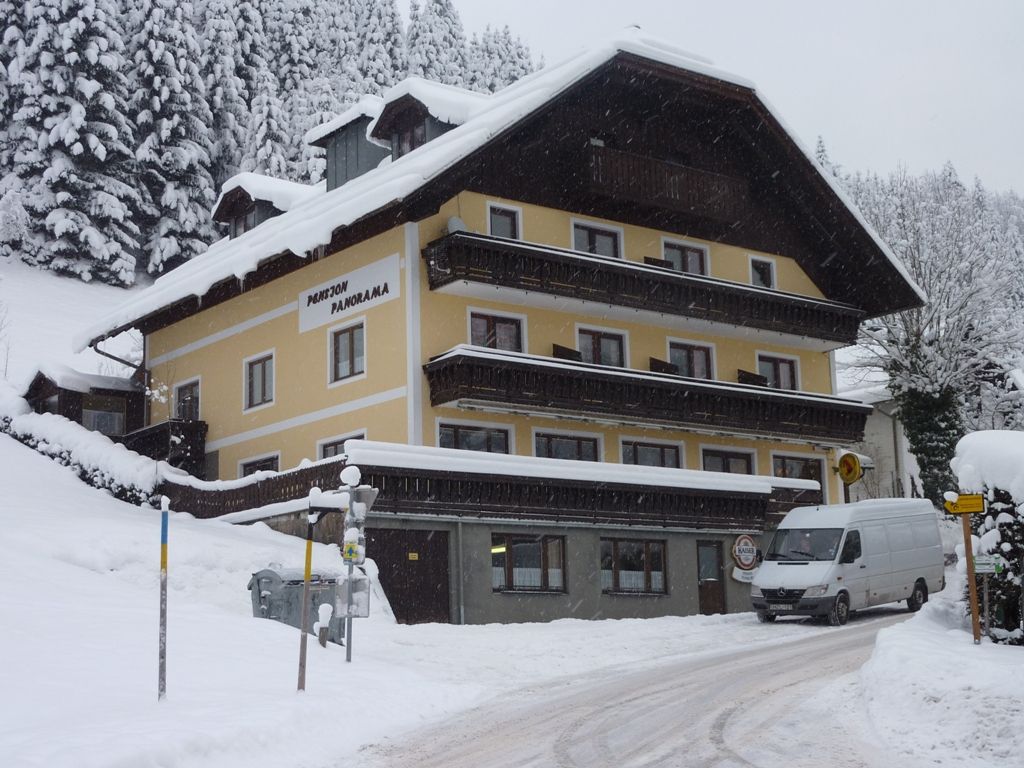Ein verschneites Gebäude mit der Aufschrift 'Pension Panorama' in einer winterlichen Landschaft.