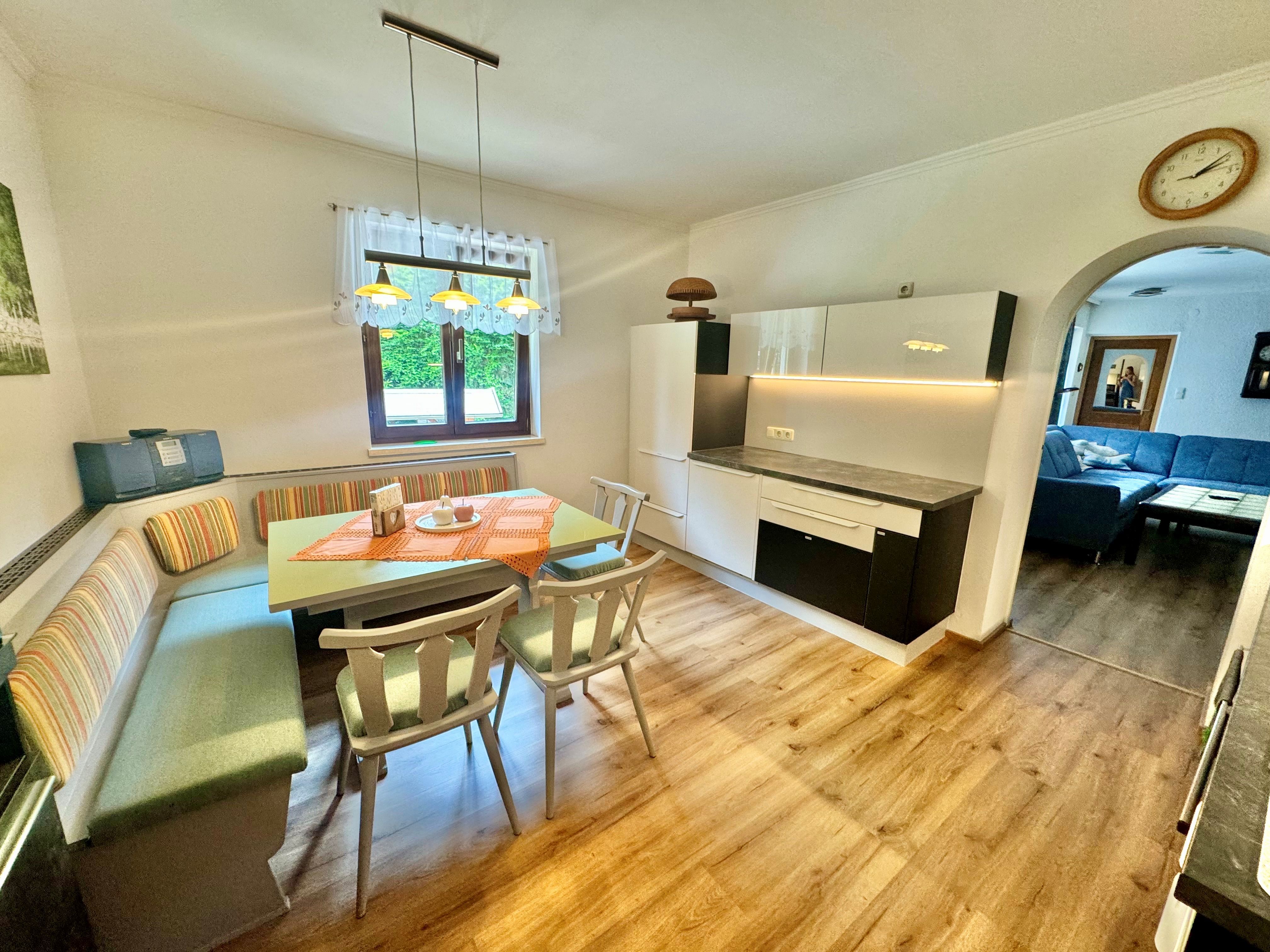 Gemütliche Küche mit Esstisch und Eckbank, daneben eine moderne Küchenzeile. Im Hintergrund ein Wohnzimmer mit blauer Couch.