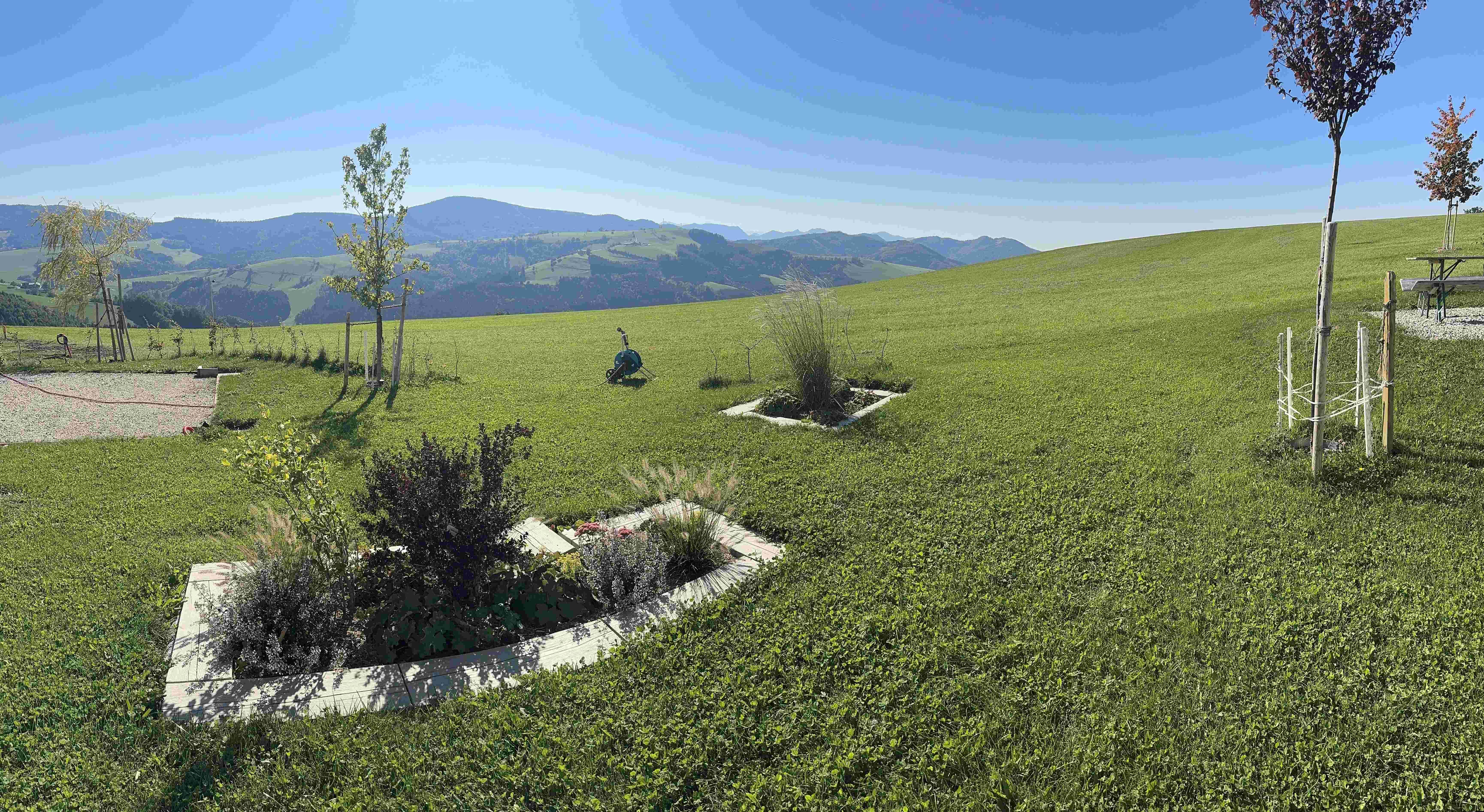 Panoramablick vom Camping am Hof Rabenberg über eine grüne Wiese mit jungen Bäumen, Blumenbeeten und weitem Panorama über die Region Moststraße