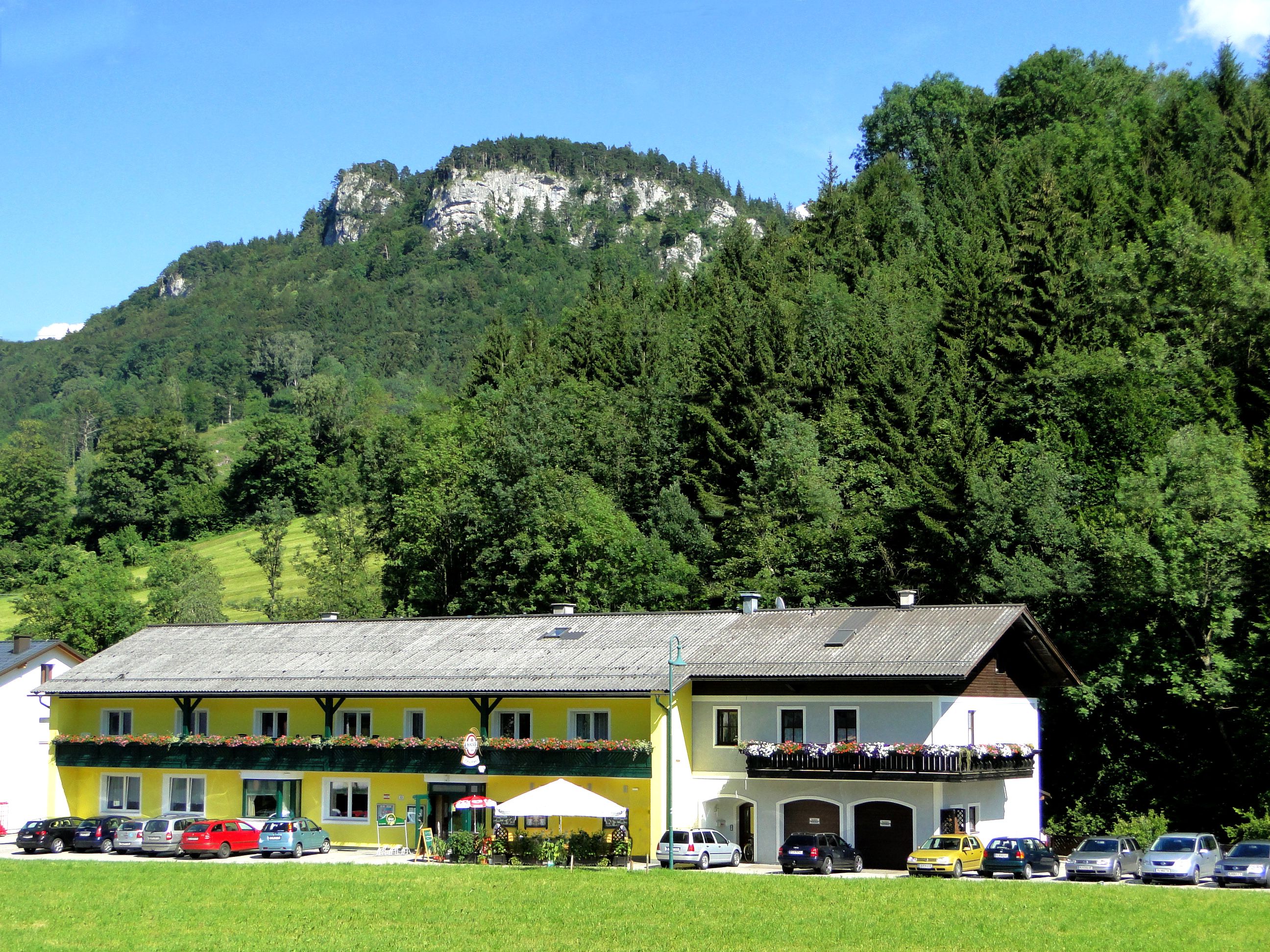Ein gelbes Gebäude mit Balkon und Blumen, umgeben von Bäumen und einem Berg im Hintergrund.