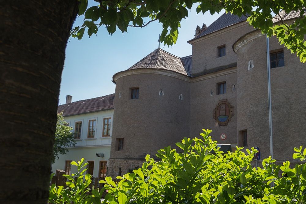Historisches Gebäude mit Rundtürmen und Wappen, umgeben von grünen Bäumen und Sträuchern.