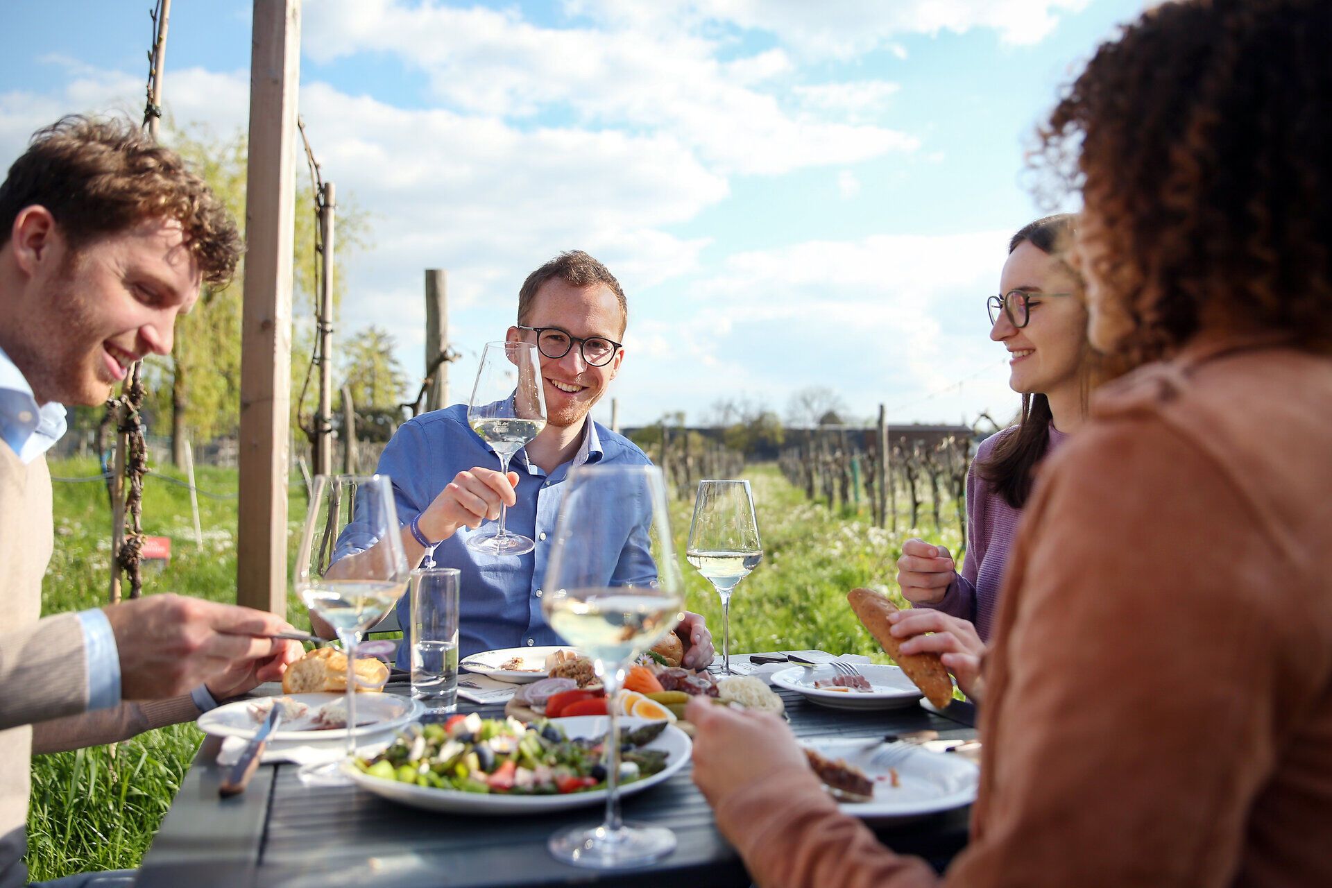Genießen Sie die entspannte Atmosphäre eines kulinarischen Erlebnisses im Freien, umgeben von sanften Weinbergen und blühenden Pflanzen. Freunde stoßen mit Gläsern an und teilen köstliche Speisen, während die Sonne sanft auf die Tafel scheint und die Landschaft in warmes Licht taucht.