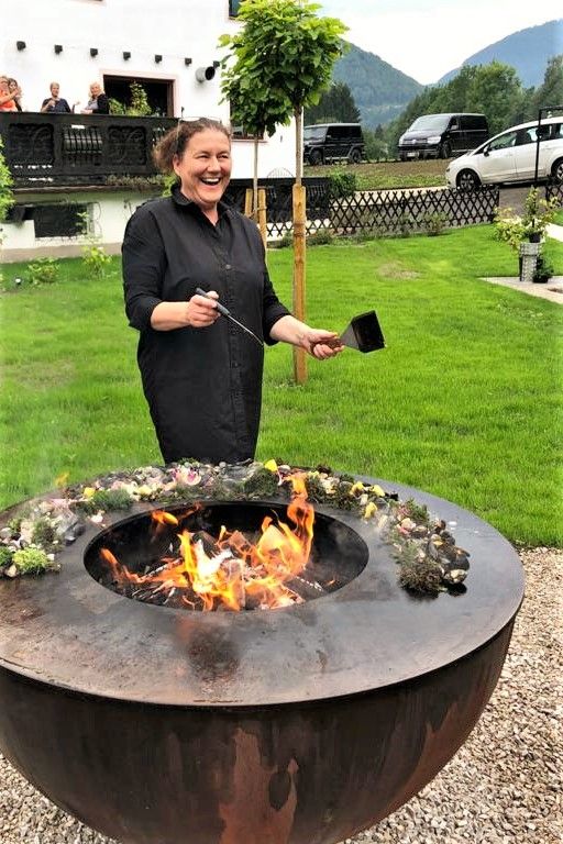 Eine Frau steht lachend an einer Feuerstelle im Freien, umgeben von grüner Landschaft und einem Gebäude im Hintergrund.