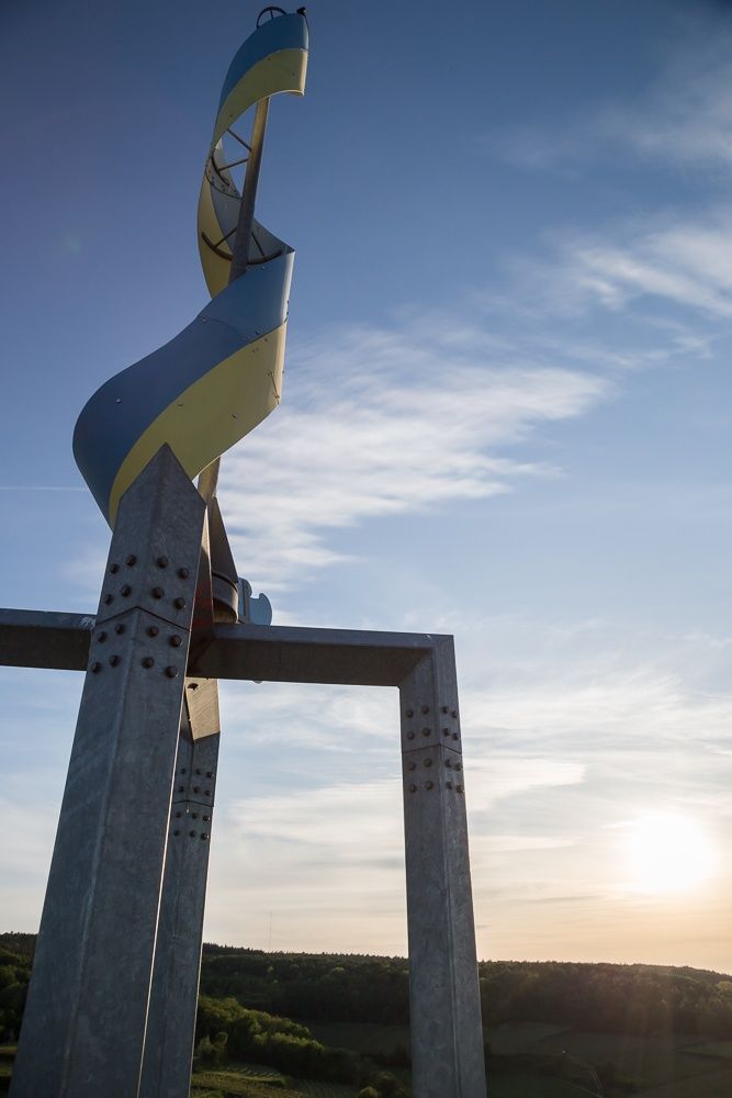 Eine große, spiralförmige Skulptur aus Metall, die an einen Korkenzieher erinnert, steht vor einem blauen Himmel mit einigen Wolken und der untergehenden Sonne.