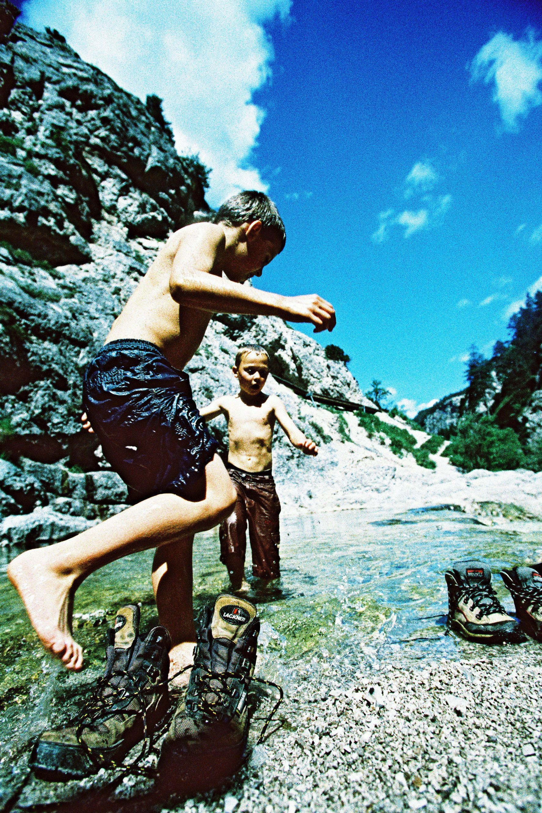 Zwei Kinder spielen im Wasser eines Gebirgsbaches, umgeben von Felsen und blauem Himmel.